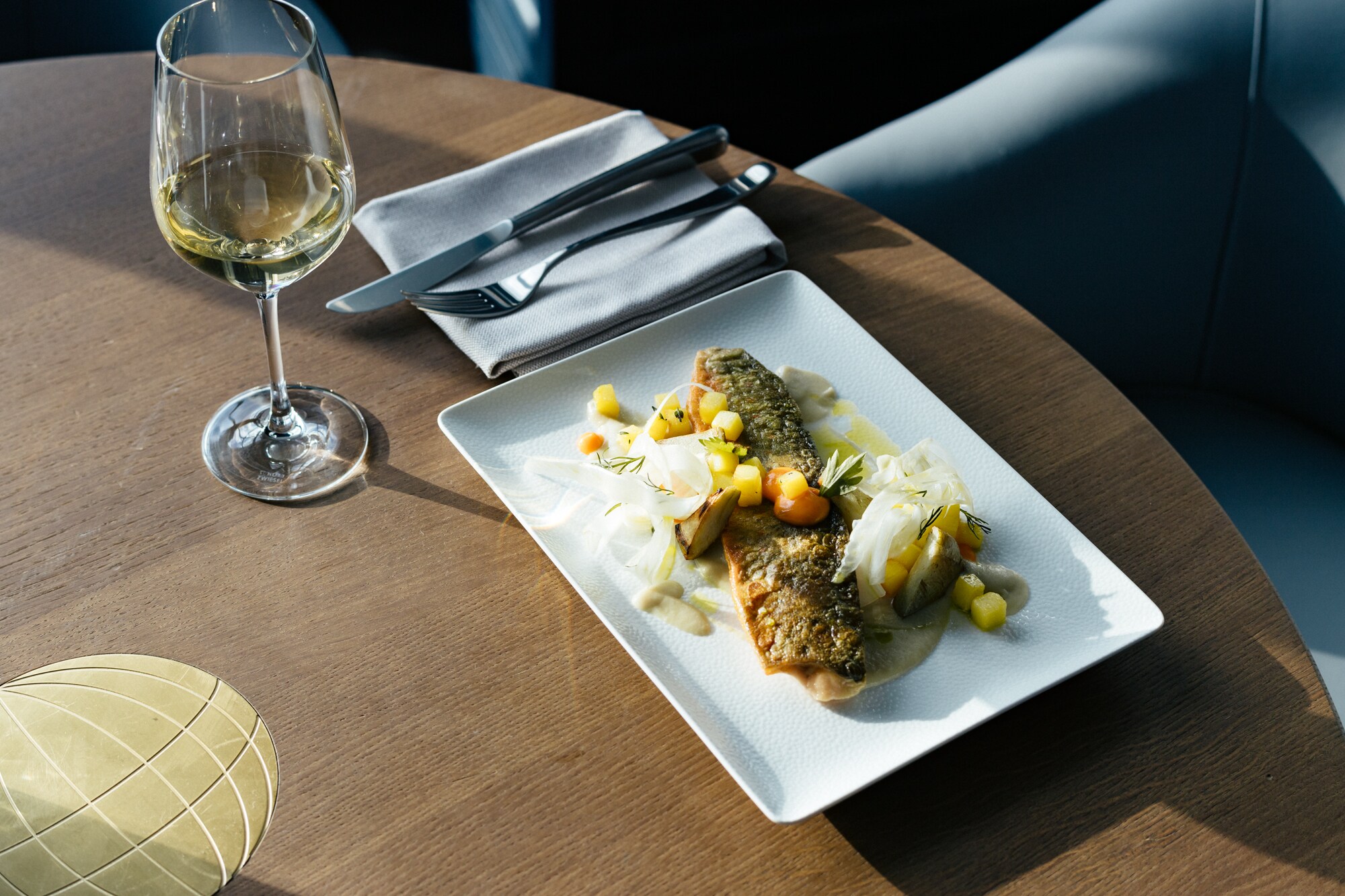 Ein Fischgericht auf einem rechteckigen Teller serviert steht auf einem Holztisch, dazu ein Glas Weißwein