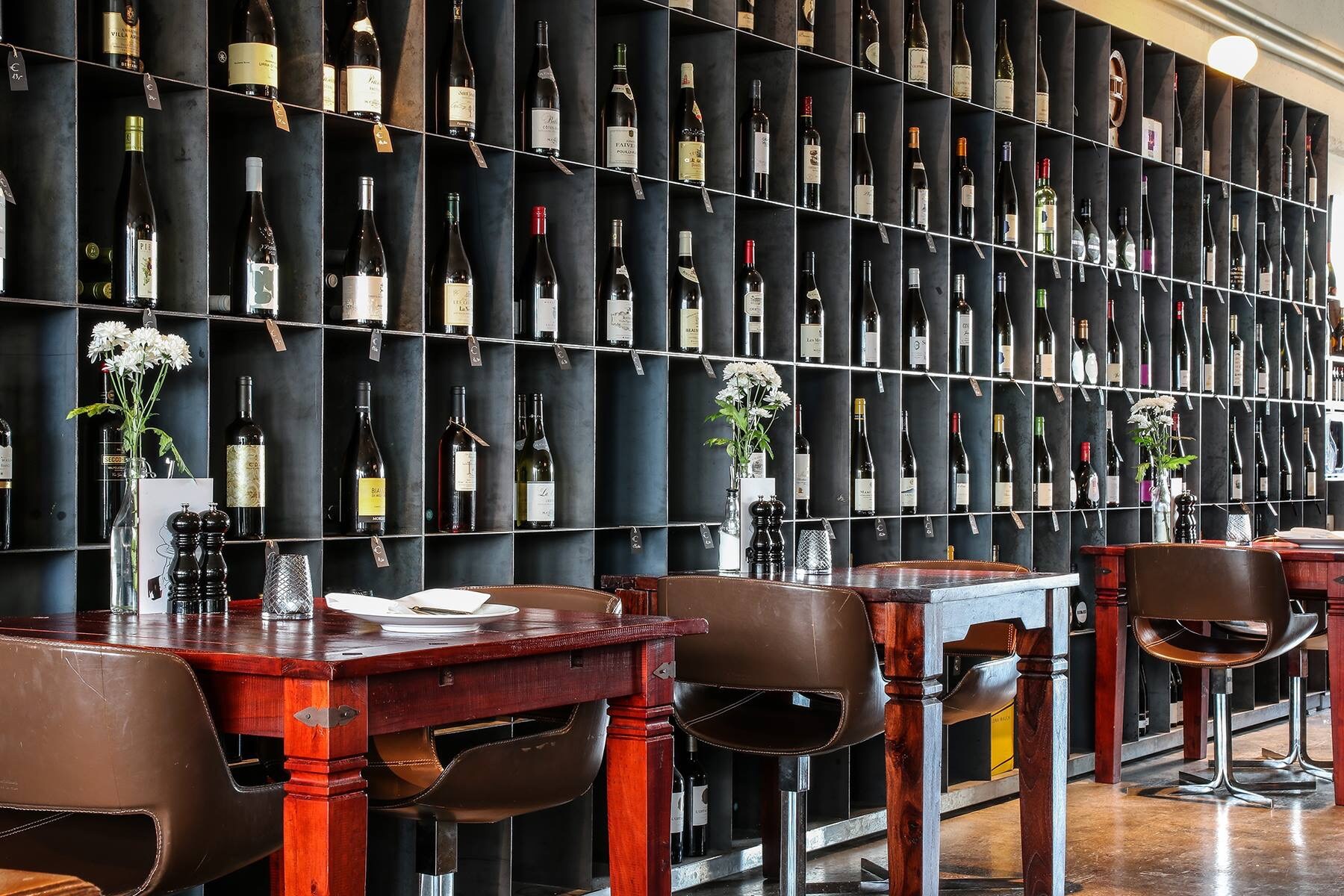 Blick auf eine Wand mit unzähligen Weinflaschen, davor stehen Holztische für zwei Personen