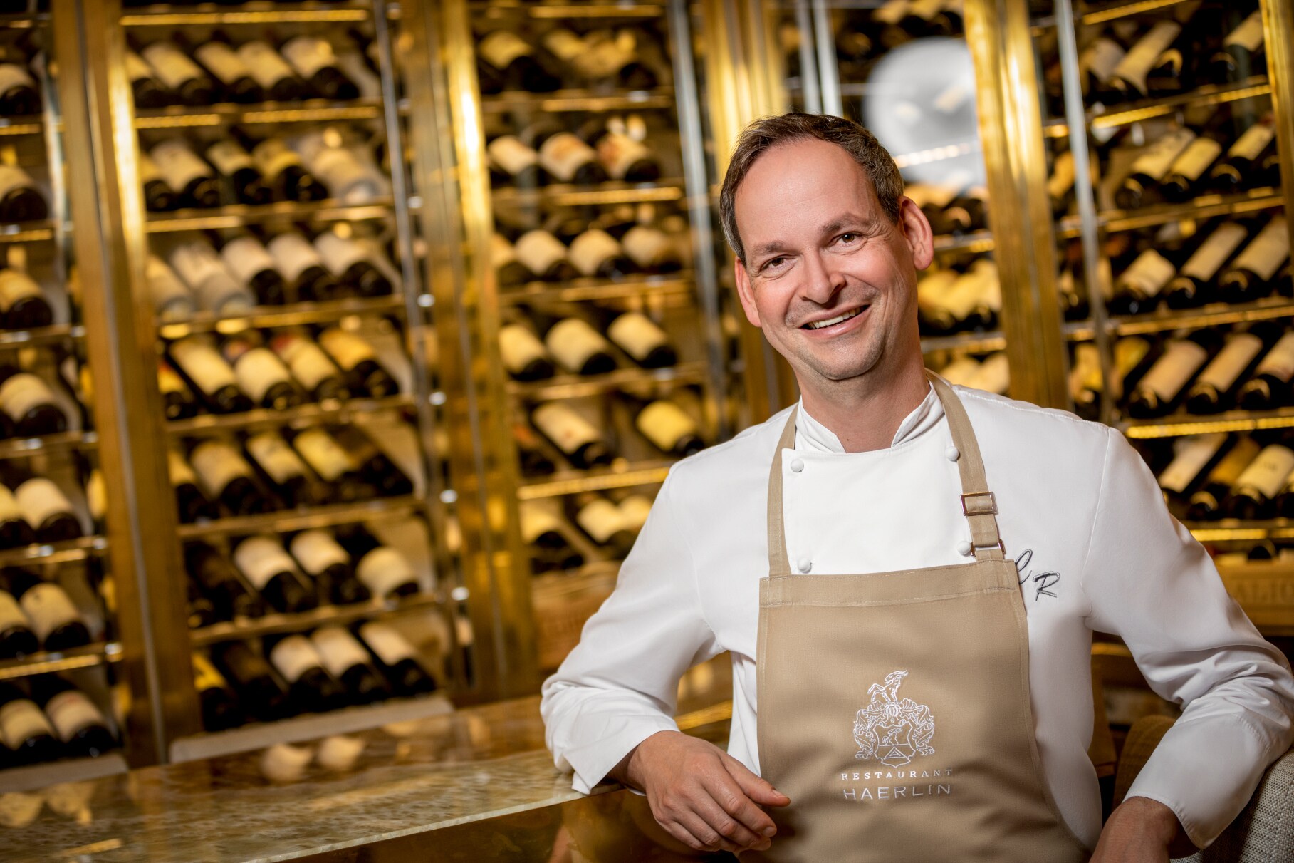 Koch mit brauner Schürze vor einen Wand voller Weinflaschen