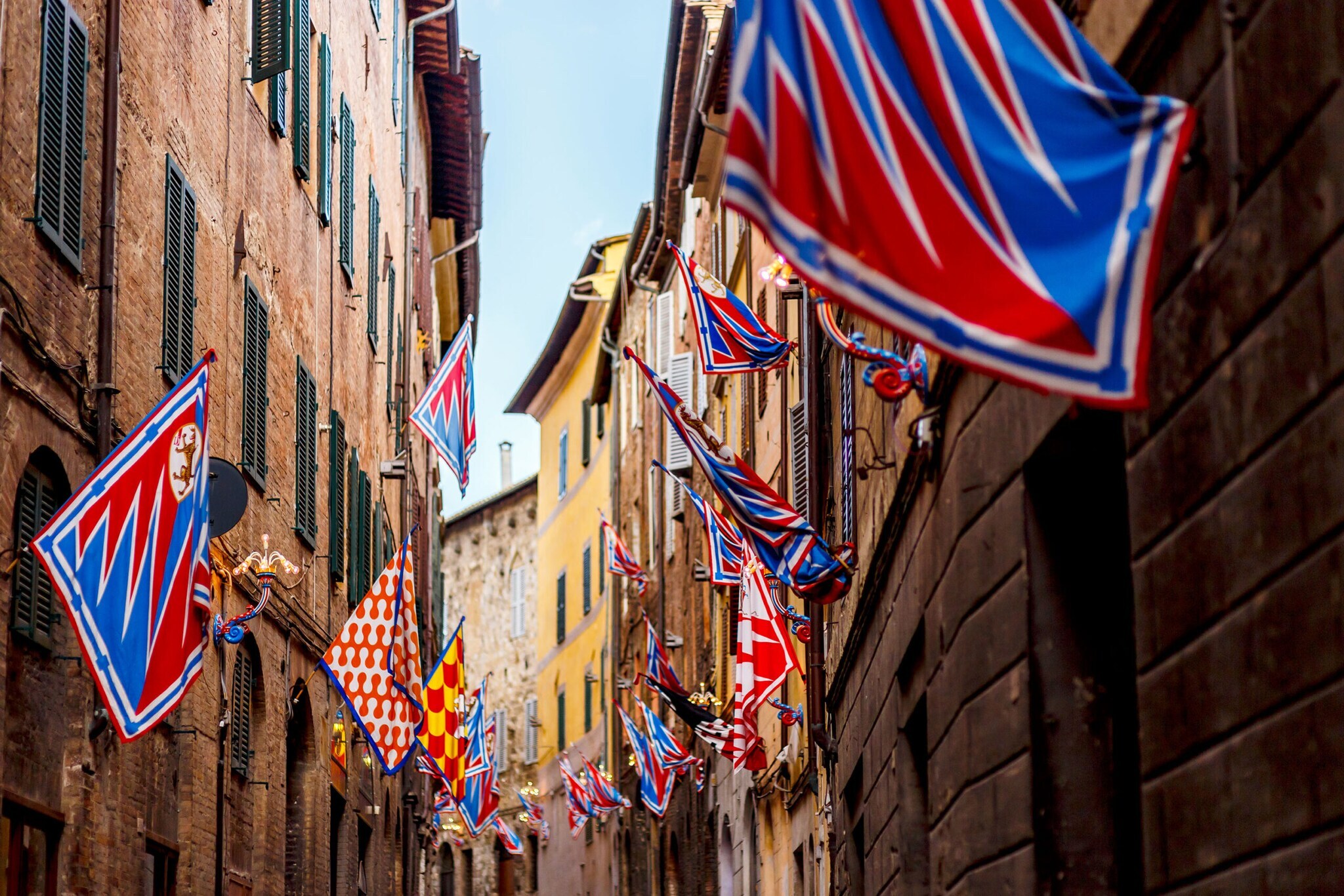 Bunte Banner mit Wappen wehen an Häuserfronten in einer Gasse beim Palio Fest in Siena