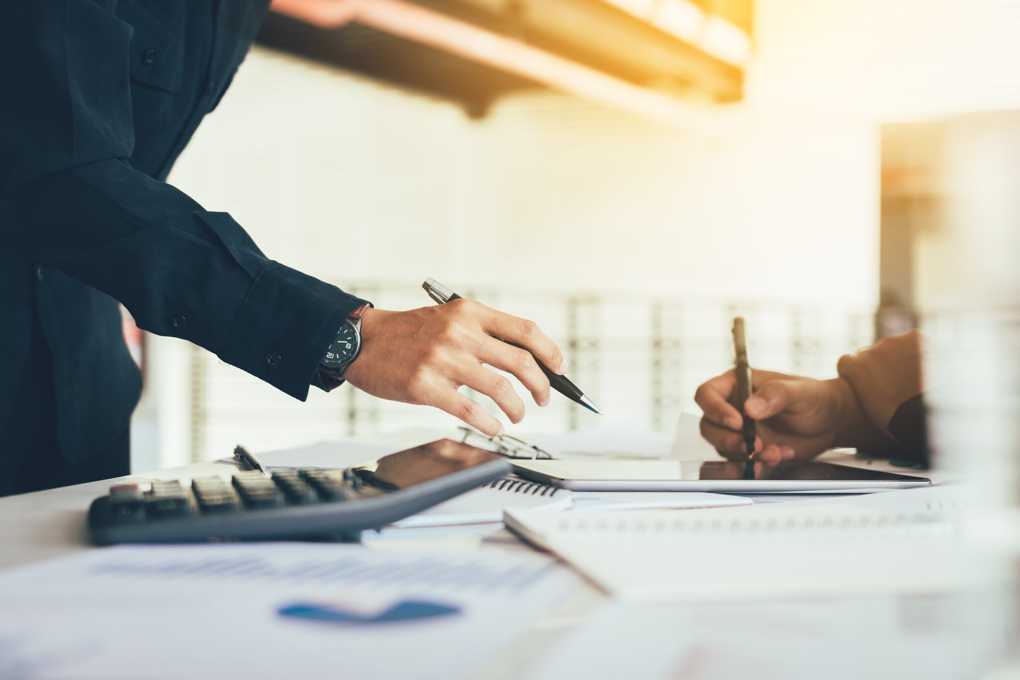 Nahansicht zweier Hände, die mit Stiften, einem Taschenrechner und Notizbüchern auf einem Tisch arbeiten