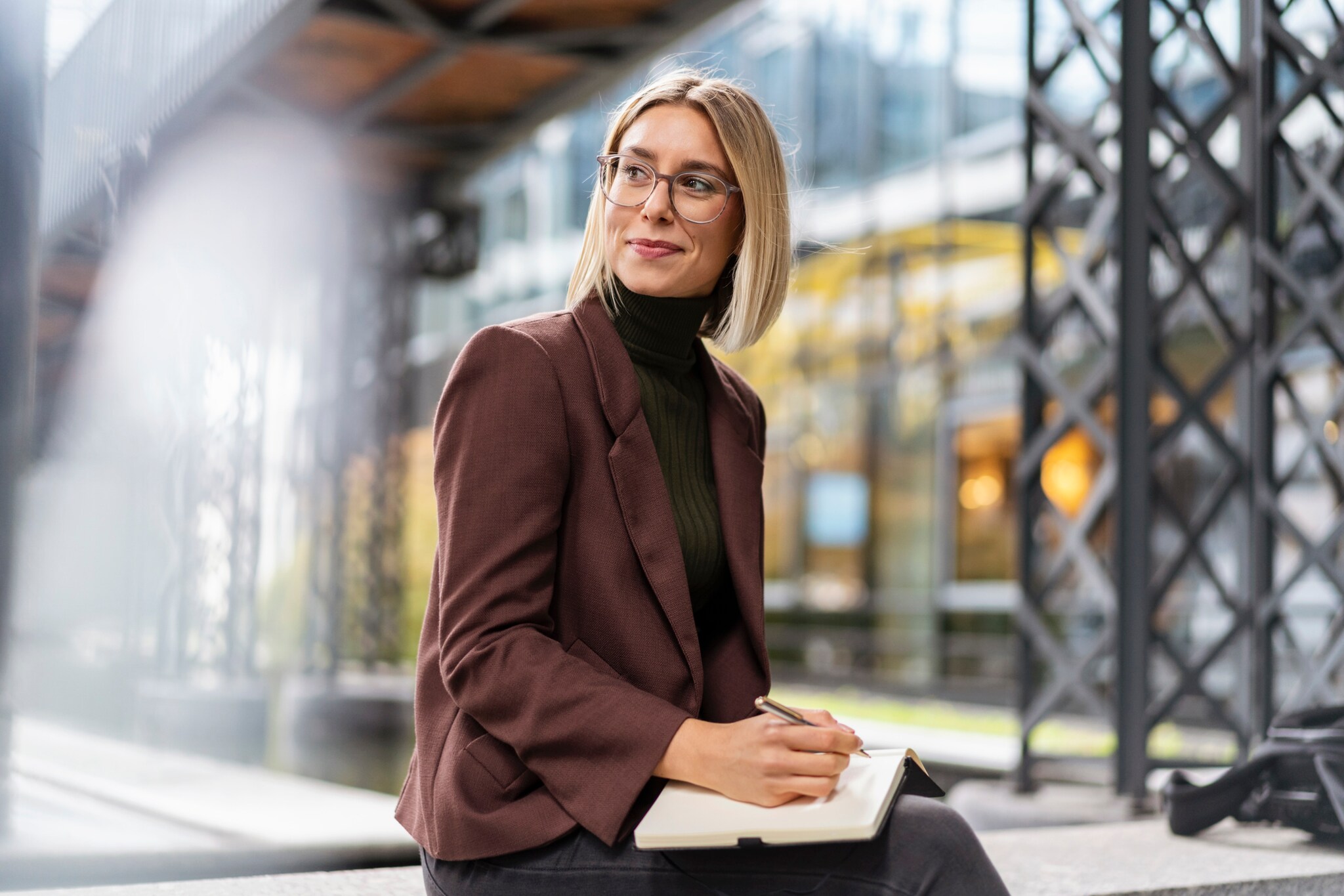 Eine Frau im Blazer sitzt mit einem Notizbuch und einem Kugelschreiber in einer industriell anmutenden Umgebung