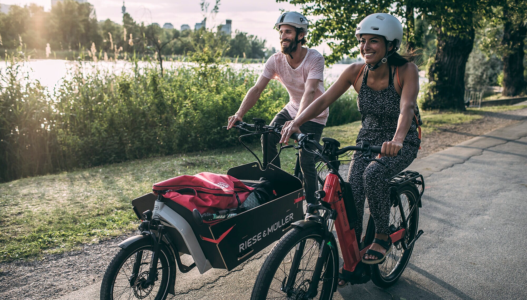 Zwei Radfahrer an einem Fluss, einer fährt ein E-Lastenrad Zwei Radfahrer an einem Fluss, einer fährt ein E-Lastenrad