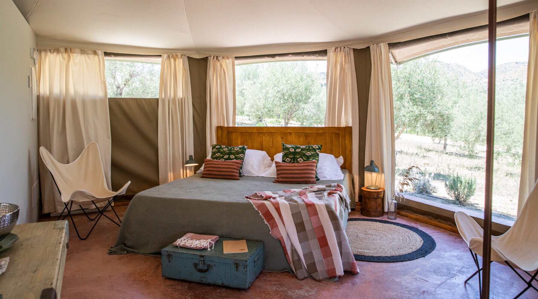 Bett in einer hellen Zelt-Lodge mit großen Fenstern, durch die Bäume und Hügel zu sehen sind