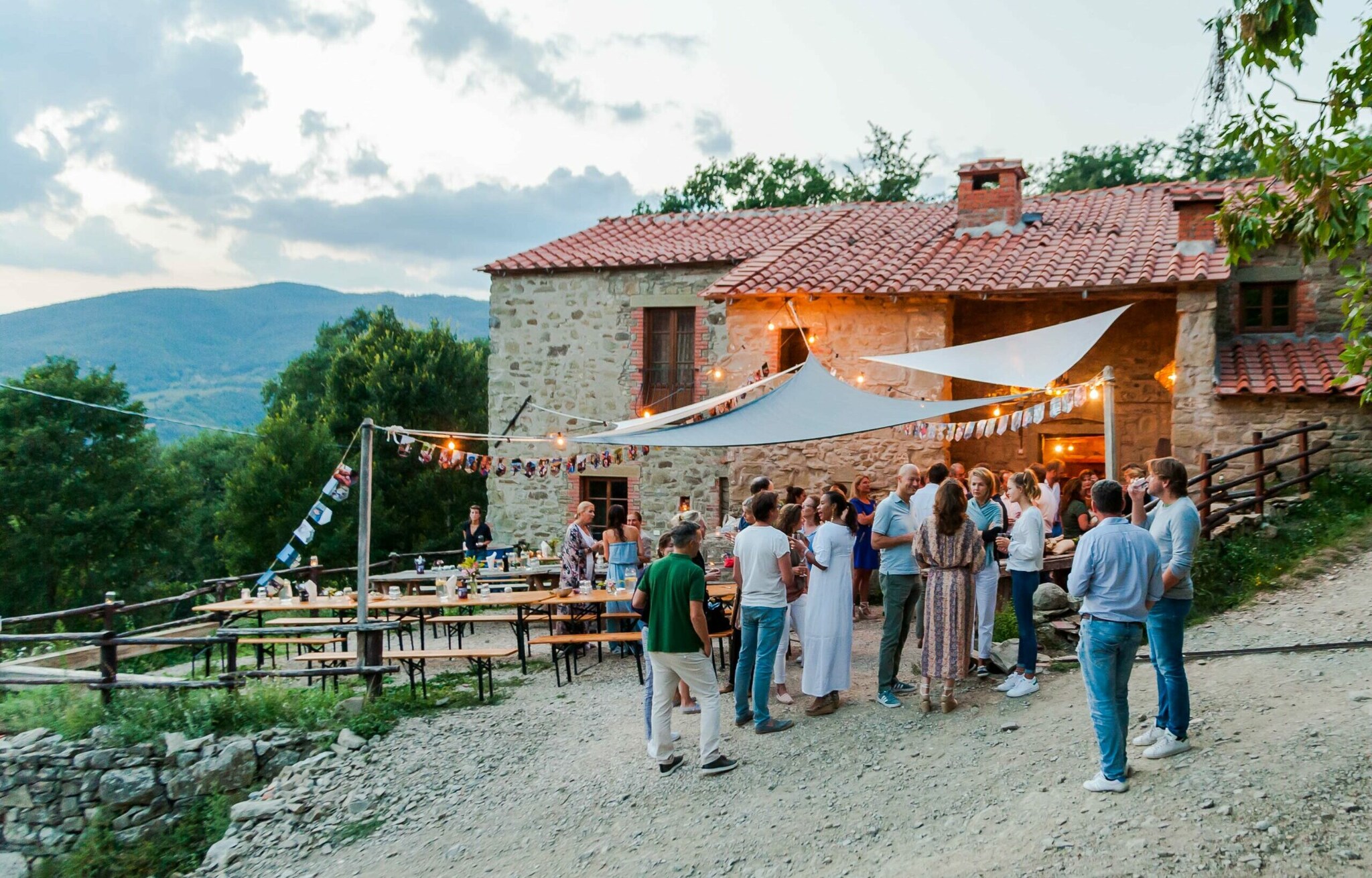 Mehrere Personen stehen um Biertische vor einem Steinhaus, im Hintergrund Berge