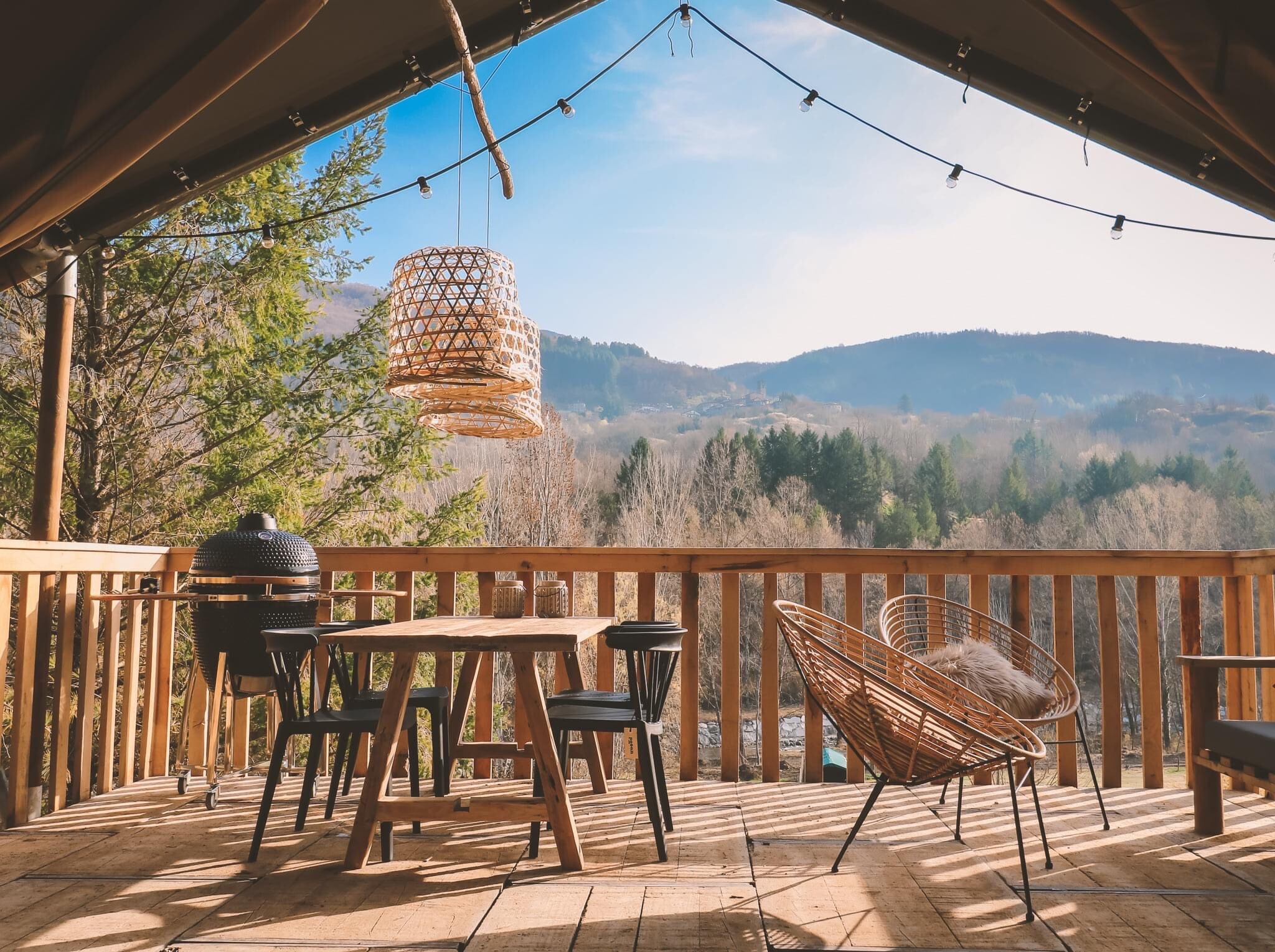 Holzterrasse mit Tisch, Stühlen und Lampen, im Hintergrund bewaldete Hügel