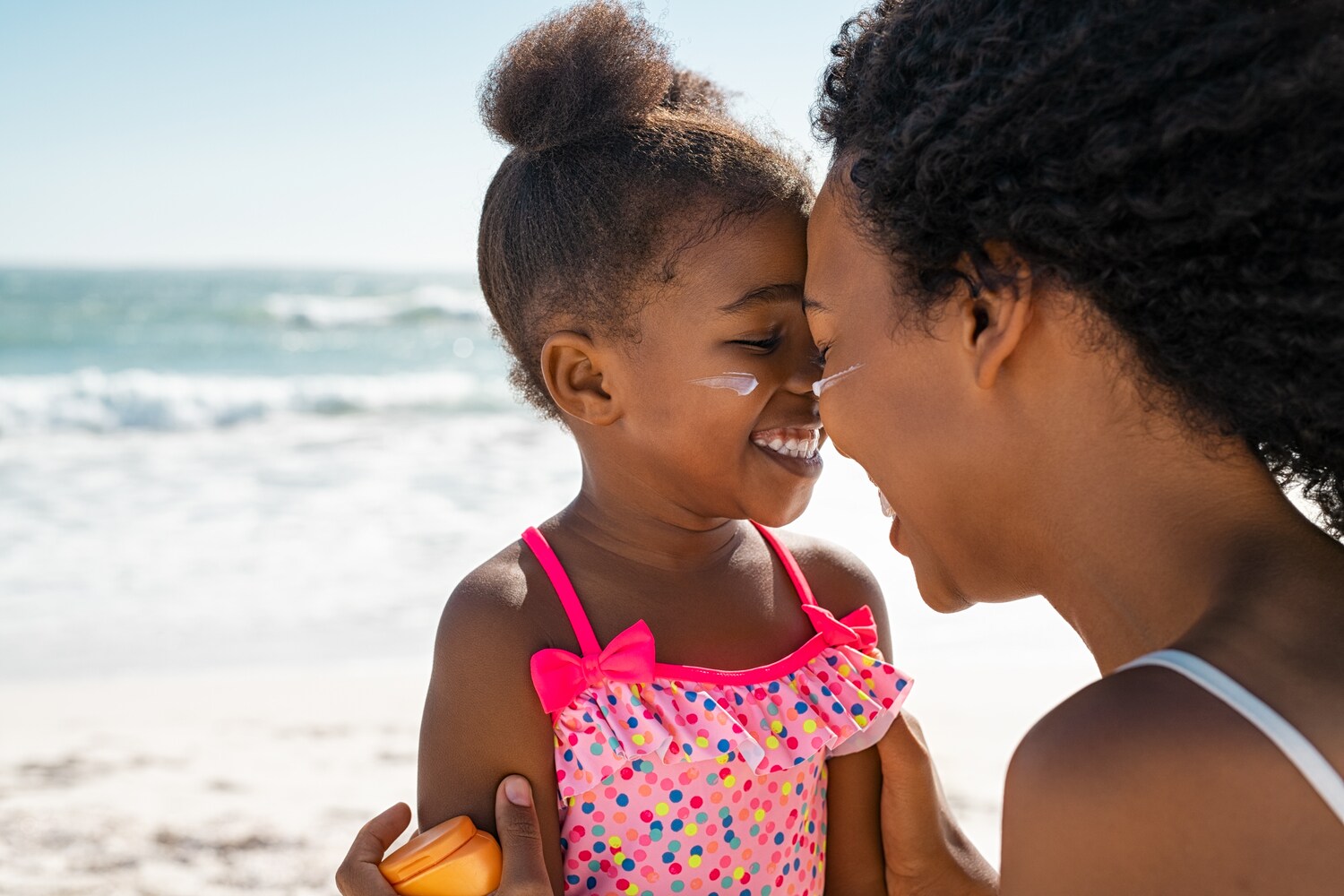 Ein kleines Mädchen am Strand mit Sonnencreme im Gesicht wird lachend von seiner Mutter umarmt