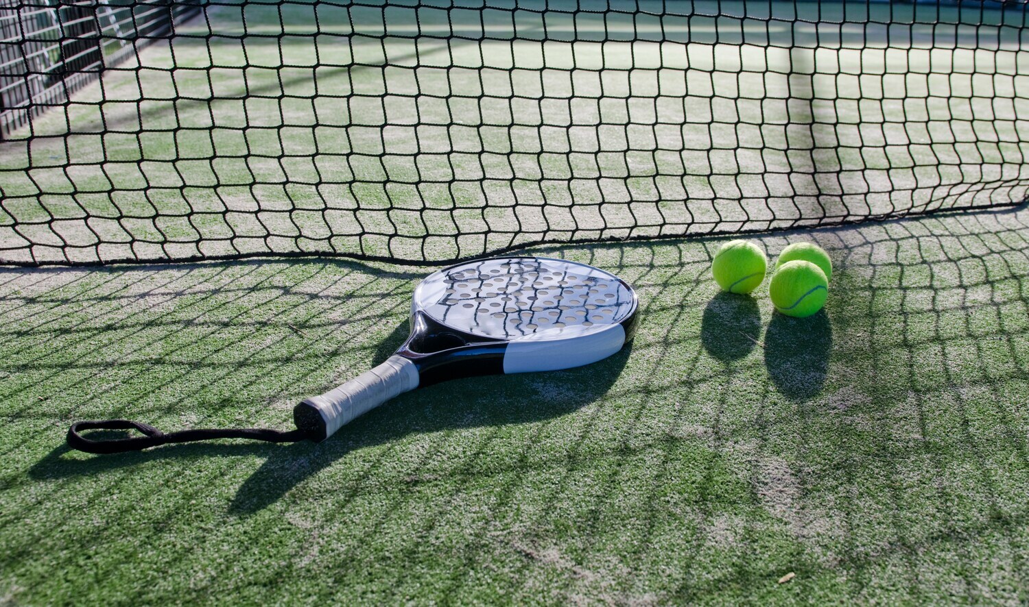Padel-Schläger mit Löchern und Schlaufe, daneben liegen drei Tennisbälle.
