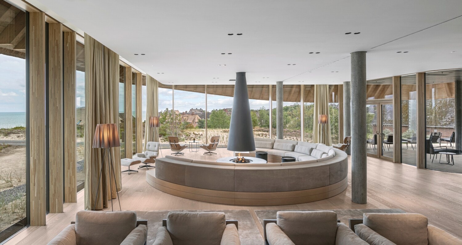 Heller Aufenthaltsraum eines Hotels in den Dünen mit Meerblick mit gerundeter Sitzgruppe um einen Kamin Heller Aufenthaltsraum eines Hotels in den Dünen mit Meerblick mit gerundeter Sitzgruppe um einen Kamin