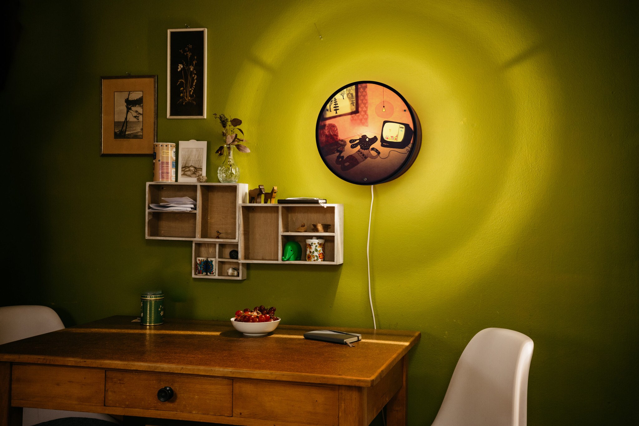 Ausschnitt einer Küche mit grüner Wand, Tisch, zwei Stühlen, ein paar kleinen Regalen an der Wand und einer Wandlampe.