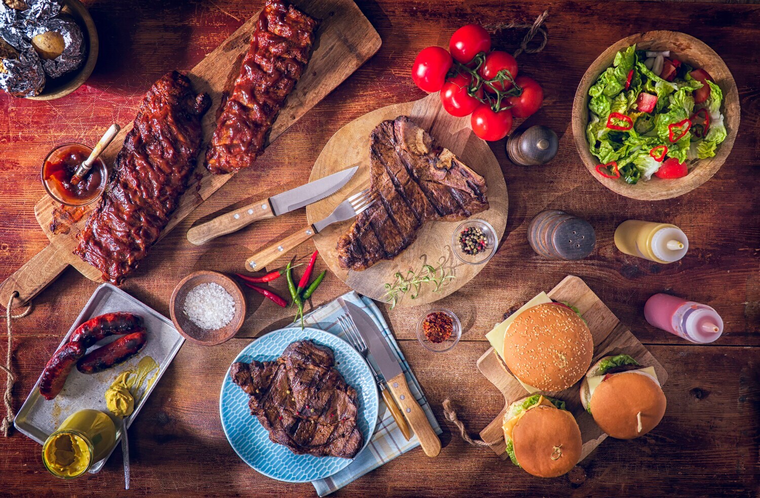 Aufsicht eines Holztisches mit gegrillten BBQ Variationen mit Steaks, Spareribs und Burgern Aufsicht eines Holztisches mit gegrillten BBQ Variationen mit Steaks, Spareribs und Burgern