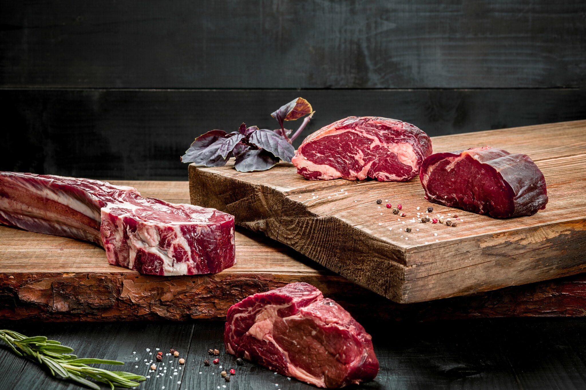 Rohes Rindfleisch auf Schneidebrett mit Gewürzen, frischem Basilikum und einem Zweig Rosmarin auf Holzgrund Rohes Rindfleisch auf Schneidebrett mit Gewürzen, frischem Basilikum und einem Zweig Rosmarin auf Holzgrund