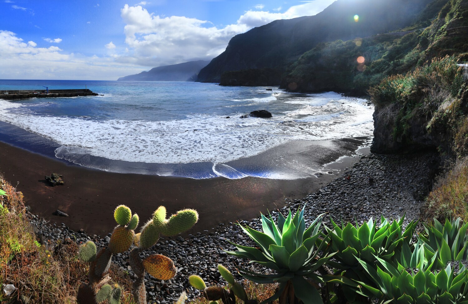 Panorama eines menschenleeren Strandes mit dunklem Sand und Steinen, umgeben von Felsen und tropischer Vegetation