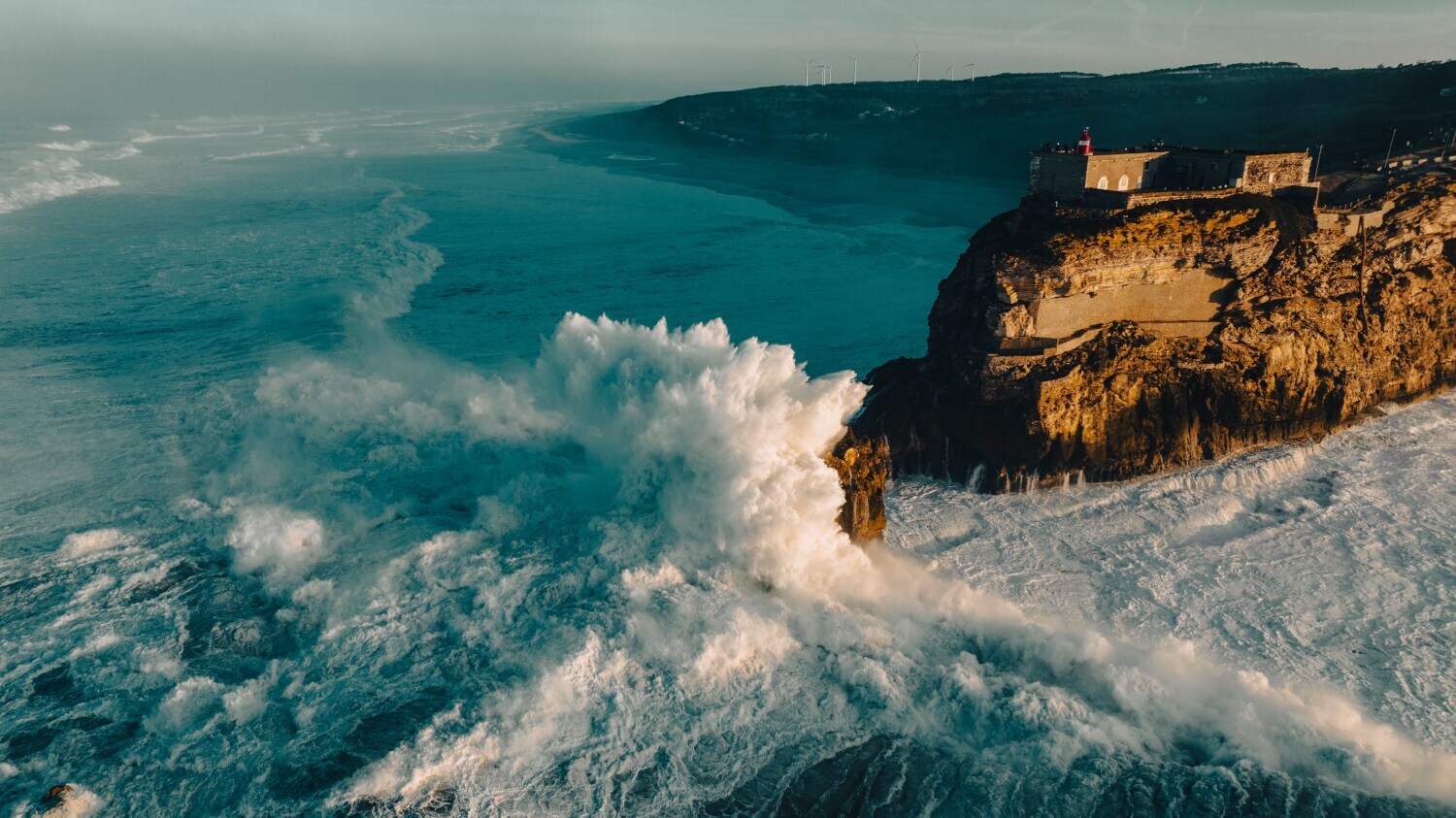 Luftaufnahme eines Küstenabschnitts mit Felsvorsprung, an dem hohe Wellen brechen