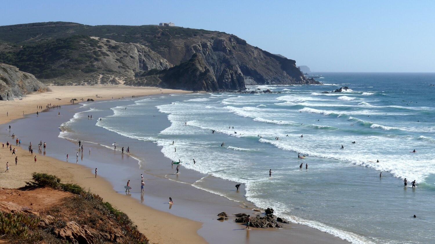 Panorama einer Bucht mit Sandstrand und Personen im Wasser, im Hintergrund Felsen