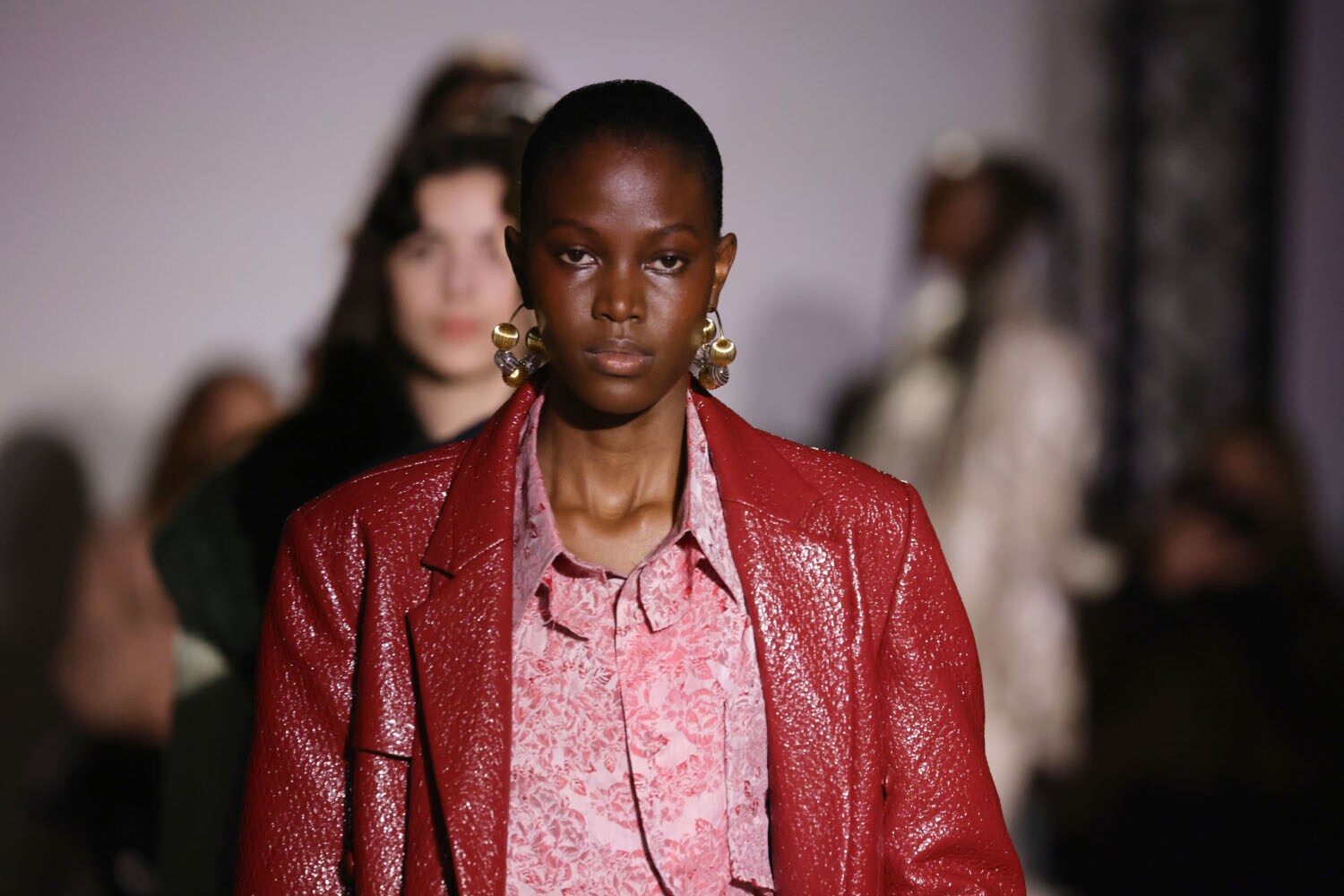 Porträt eines weiblichen, dunkelhäutigen Models mit goldenen Ohrringen und glitzernder, roter Lederjacke auf einem Laufsteg