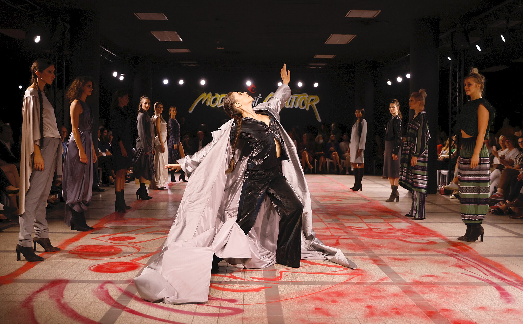 Eine Tänzerin mit weitem Mantel auf dem Catwalk umringt von stehenden Models Eine Tänzerin mit weitem Mantel auf dem Catwalk umringt von stehenden Models