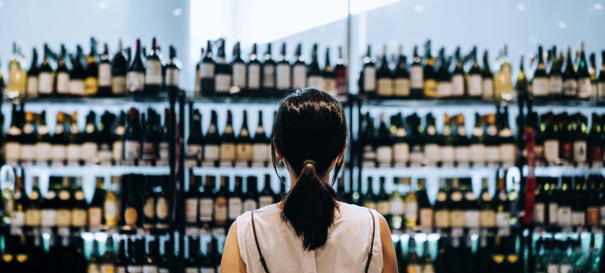 Eine dunkelhaarige Frau von hinten, die vor einem Weinregal steht.