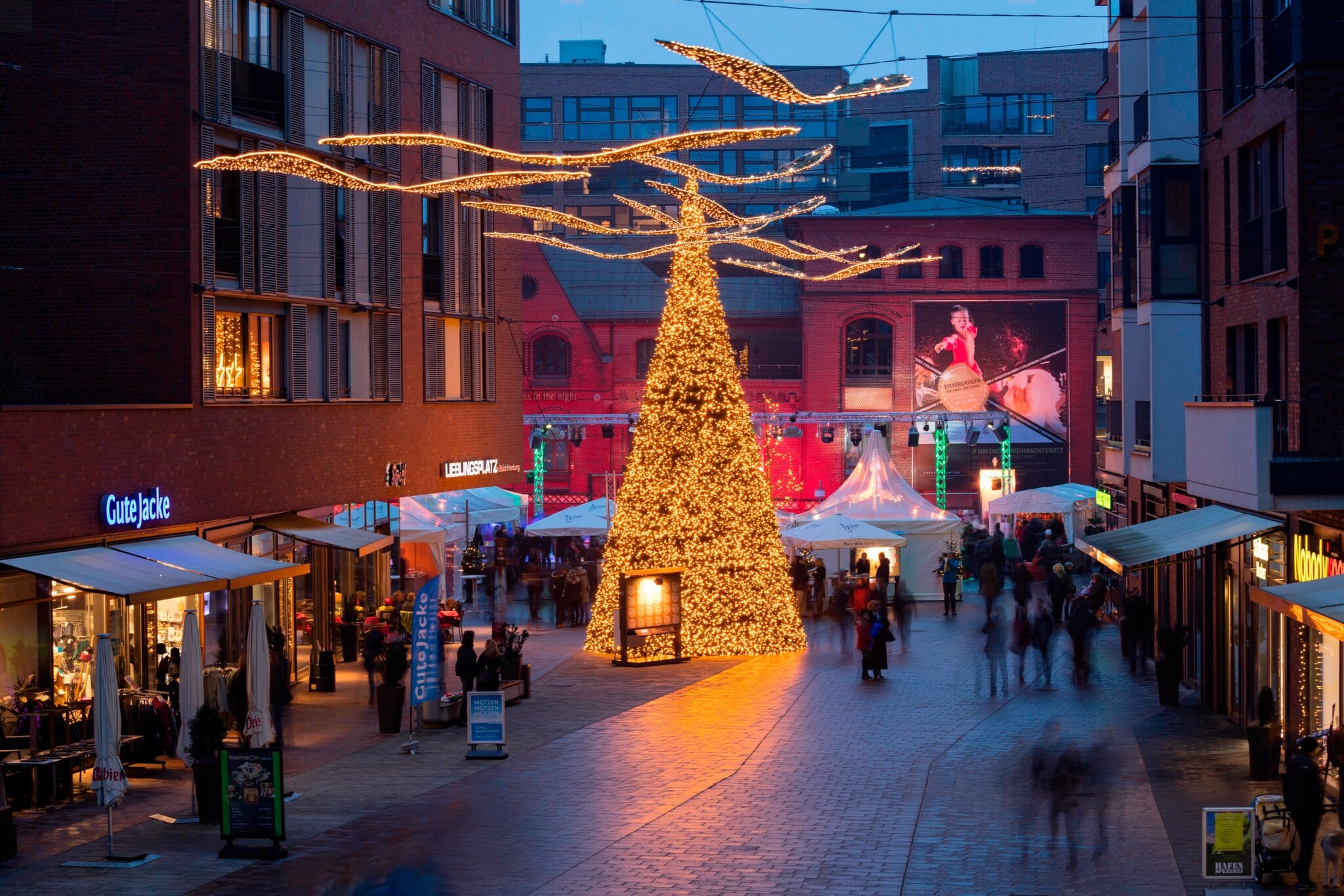 Erleuchteter Weihnachtsbaum mit Marktständen zwischen modernen Gebäuden in einer Einkaufsstraße Erleuchteter Weihnachtsbaum mit Marktständen zwischen modernen Gebäuden in einer Einkaufsstraße