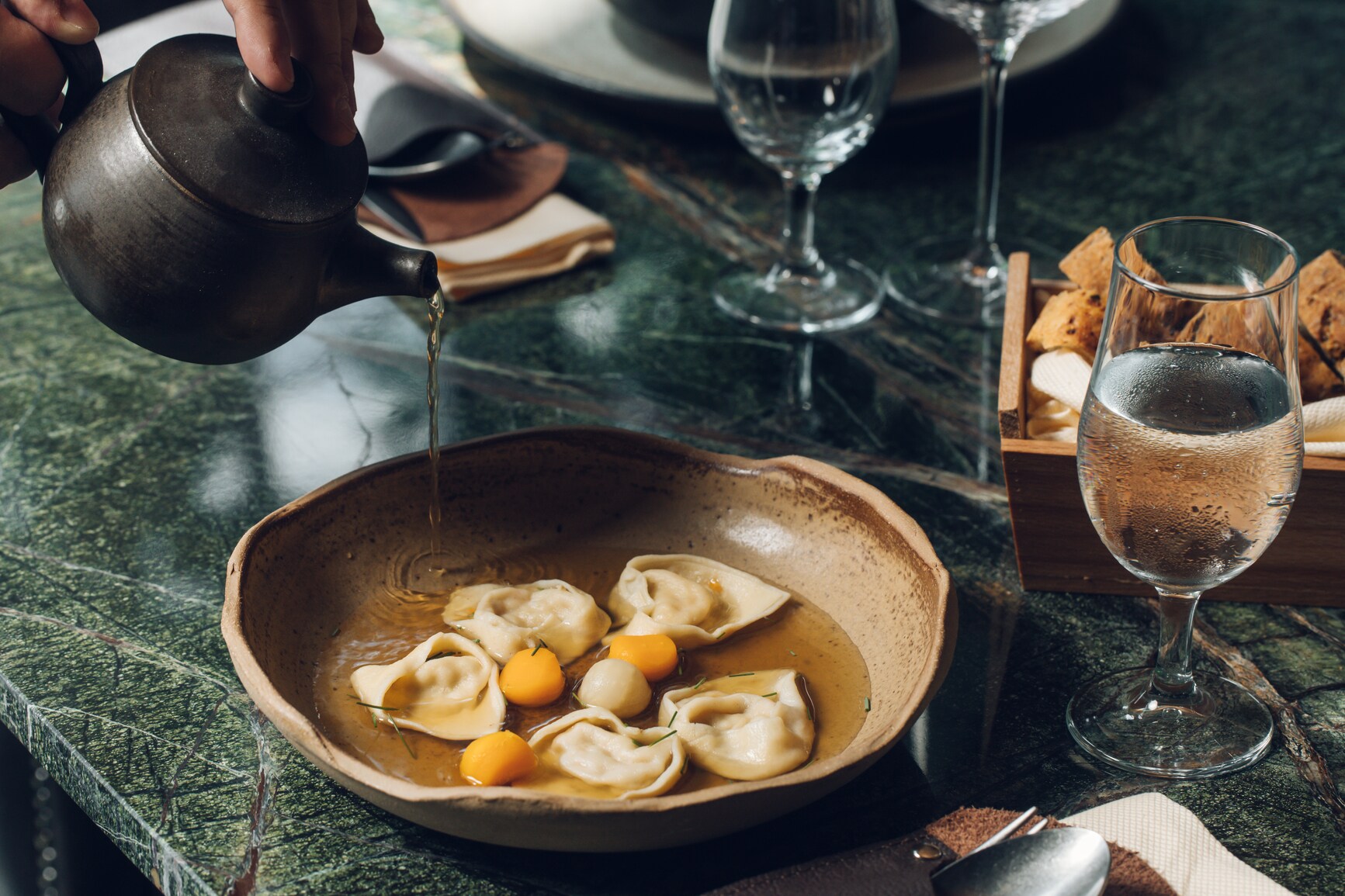 An einem stilvoll gedeckten Restauranttisch läuft Brühe aus einer Kanne in einen Teller mit gefüllter Pasta
