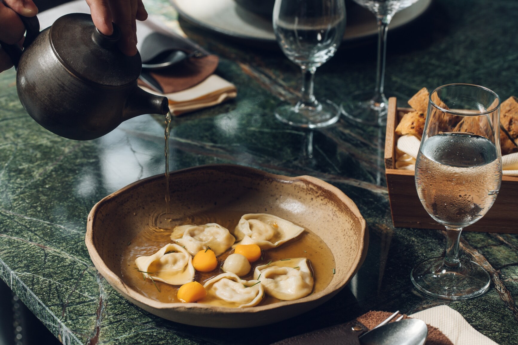 An einem stilvoll gedeckten Restauranttisch läuft Brühe aus einer Kanne in einen Teller mit gefüllter Pasta An einem stilvoll gedeckten Restauranttisch läuft Brühe aus einer Kanne in einen Teller mit gefüllter Pasta
