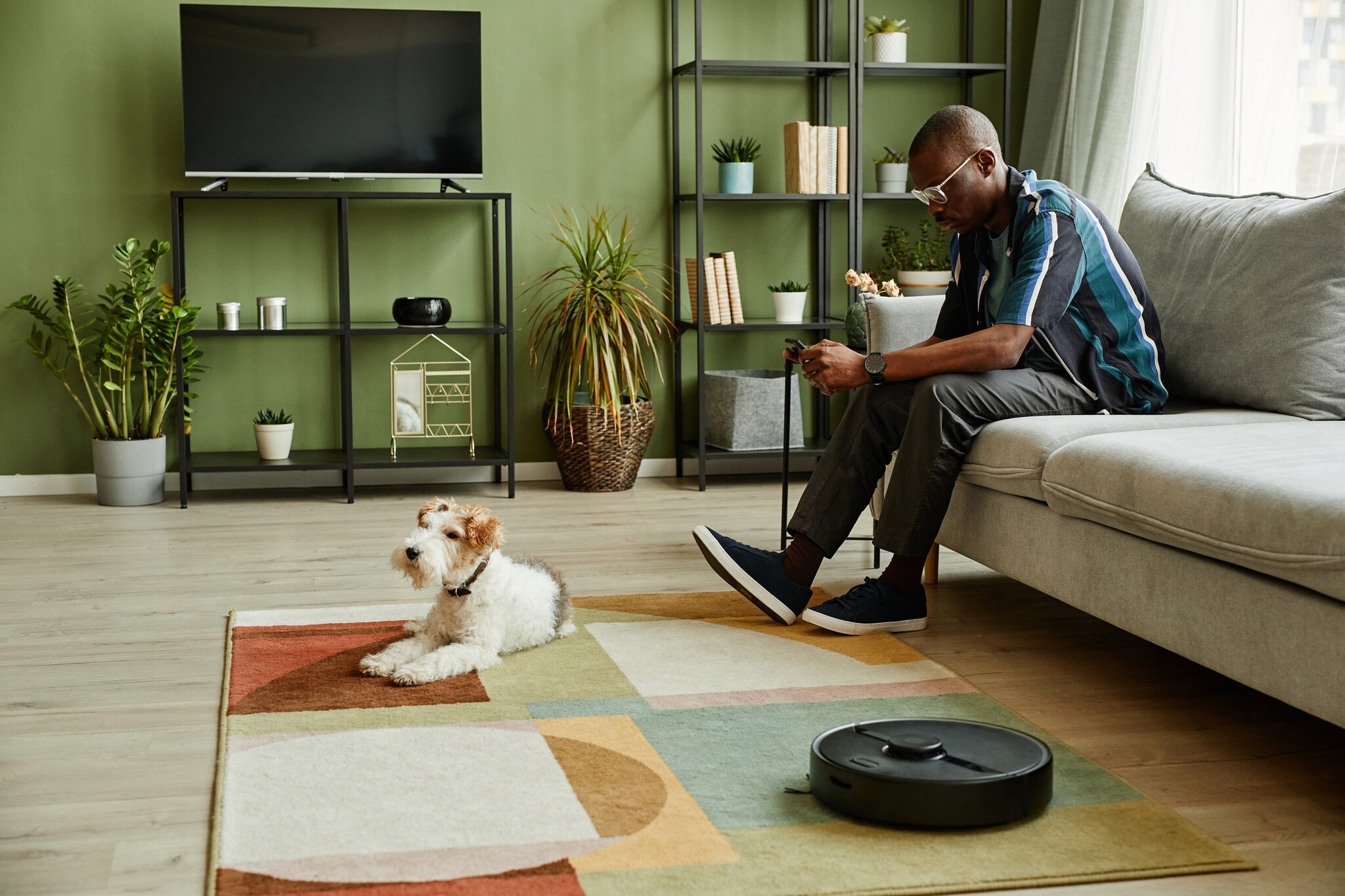 Ein Mann sitzt im Wohnzimmer auf dem Sofa und schaut auf sein Handy in der Hand. Vor ihm liegt ein Hund auf dem Teppich, daneben arbeitet ein Saugroboter. Ein Mann sitzt im Wohnzimmer auf dem Sofa und schaut auf sein Handy in der Hand. Vor ihm liegt ein Hund auf dem Teppich, daneben arbeitet ein Saugroboter.