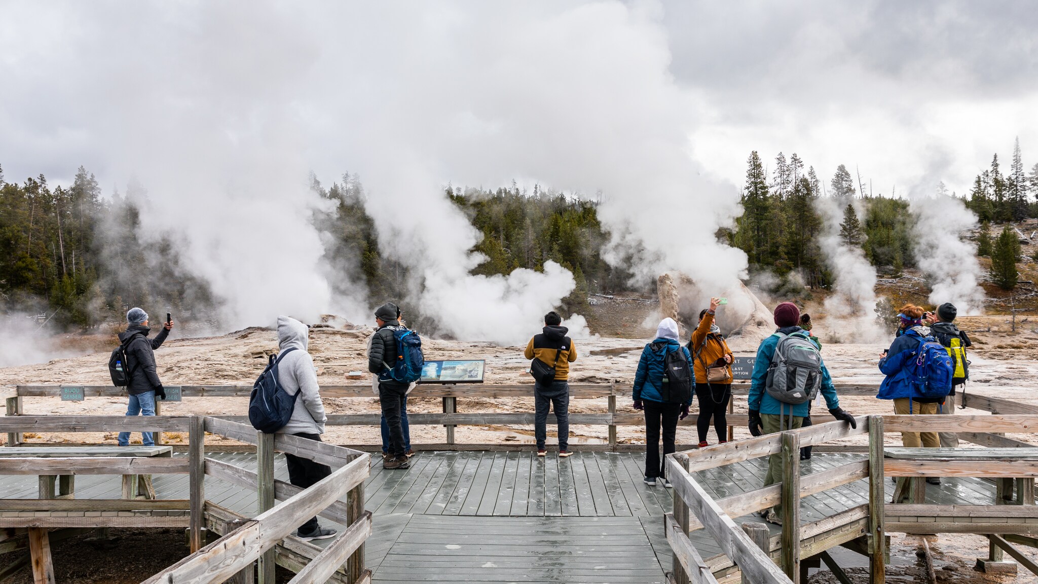 Menschen beobachten im Yellowstone-Nationalpark von einem Holzsteg aus dampfende Thermalquellen. Menschen beobachten im Yellowstone-Nationalpark von einem Holzsteg aus dampfende Thermalquellen.