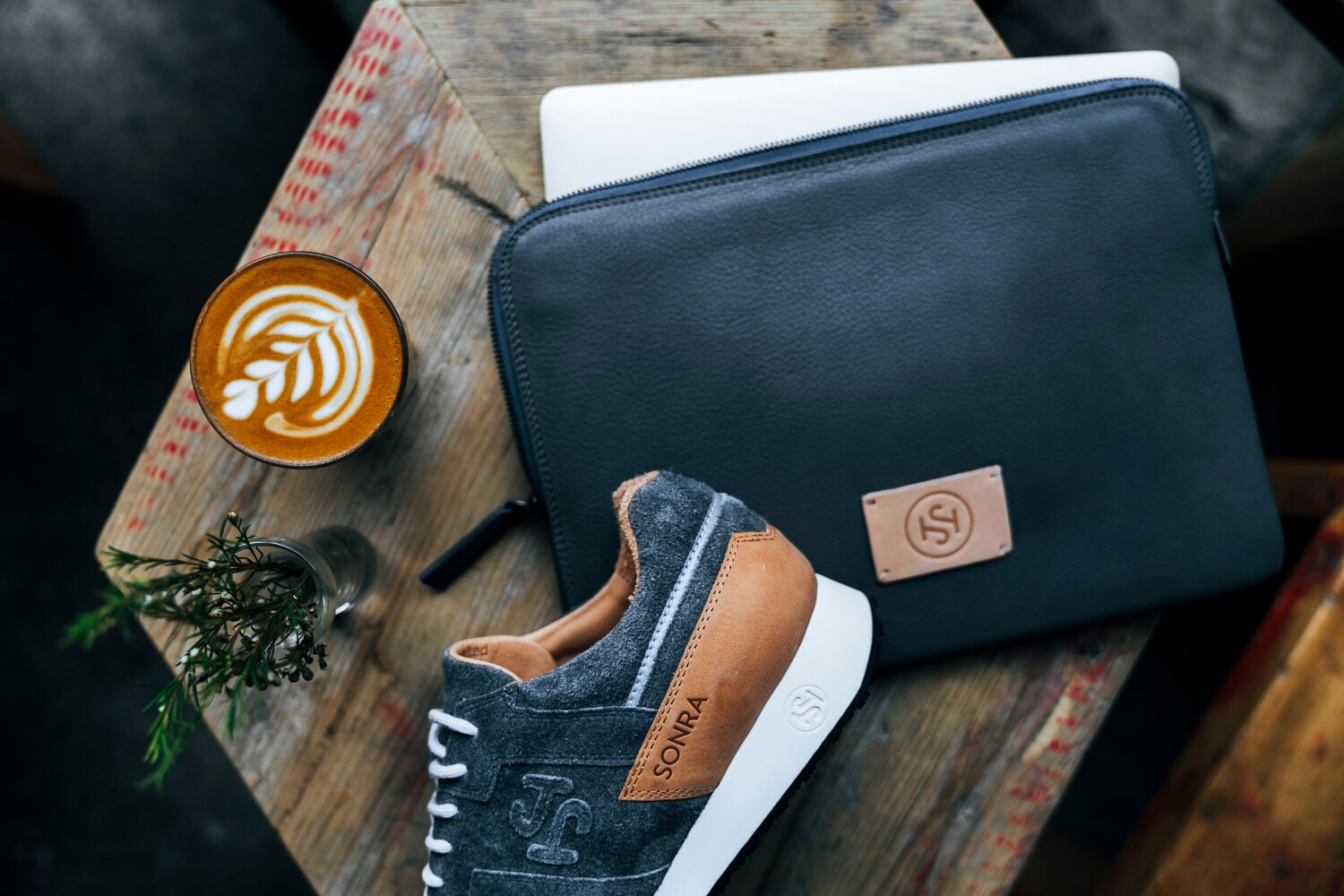 Ein Turnschuh von Sonra in Dunkelblau, Braun und Weiß auf einem Holzbrett. Daneben eine dunkelblaue Ledertasche, eine Vase und ein Kaffee. Ein Turnschuh von Sonra in Dunkelblau, Braun und Weiß auf einem Holzbrett. Daneben eine dunkelblaue Ledertasche, eine Vase und ein Kaffee.