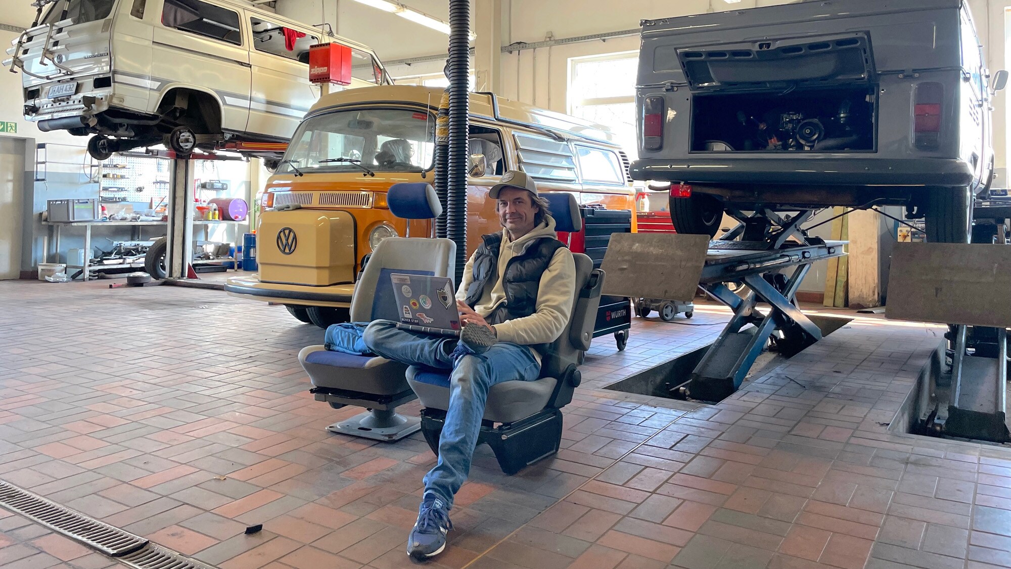 Ein Mann mit Laptop sitzt auf einem ausgebauten Fahrersitz in einer Autowerkstatt Ein Mann mit Laptop sitzt auf einem ausgebauten Fahrersitz in einer Autowerkstatt