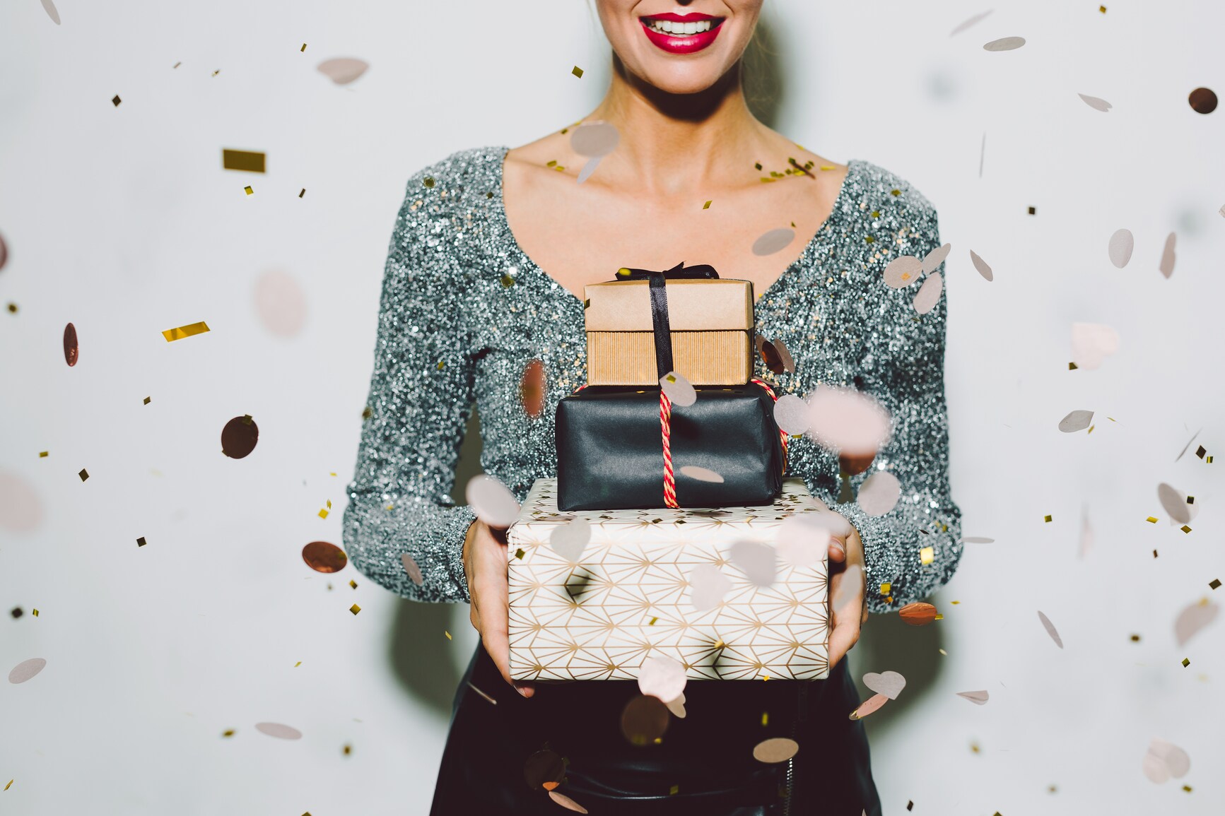 Eine Frau hält drei eingepackte Geschenke in den Händen vor ihrem Körper, um sie herum fliegt Konfetti Eine Frau hält drei eingepackte Geschenke in den Händen vor ihrem Körper, um sie herum fliegt Konfetti