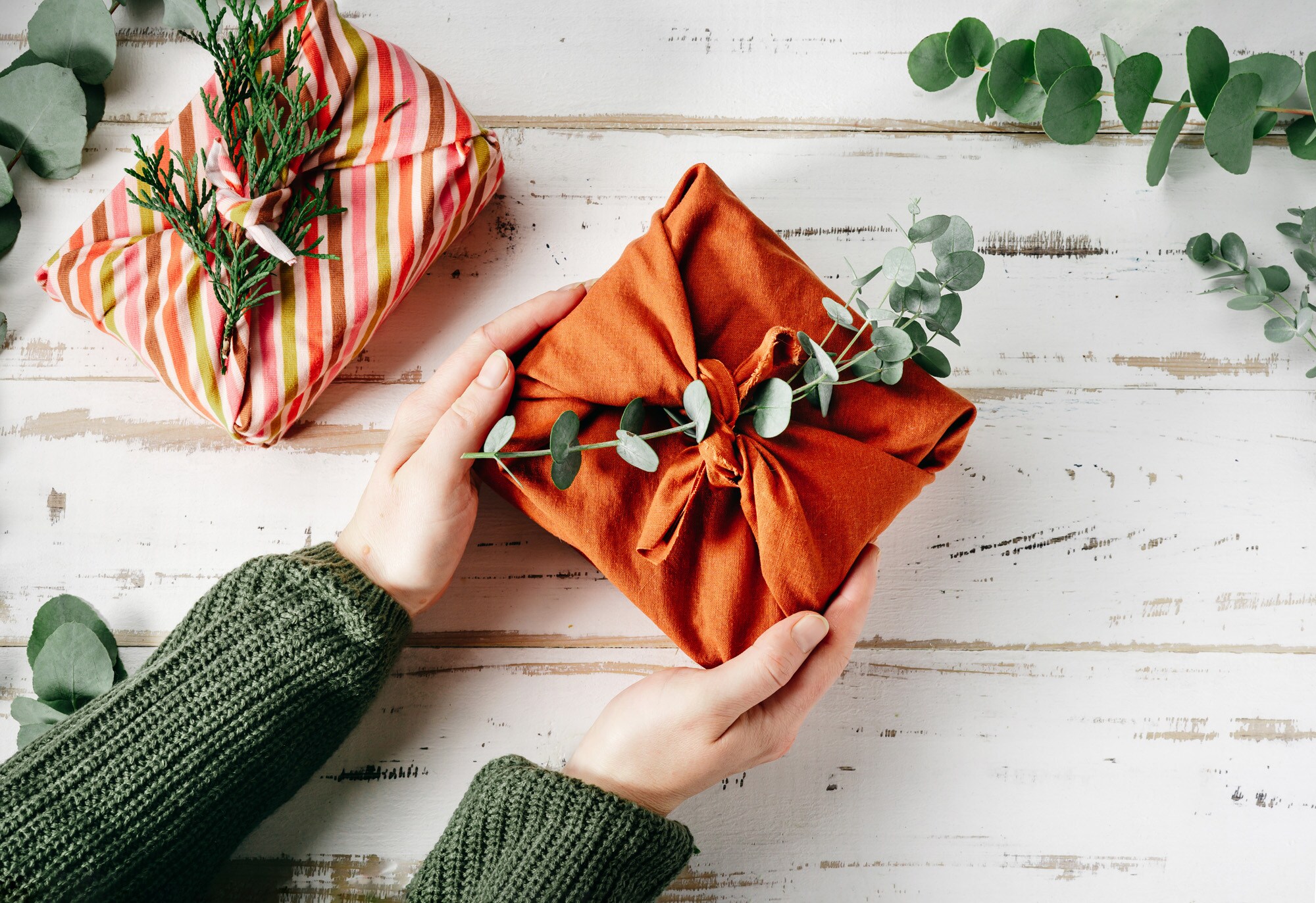Ein Geschenk, verpackt in einem orangefarbenen Tuch und geschmückt mit einem grünen Zweig, wird von zwei Händen gehalten. Ein Geschenk, verpackt in einem orangefarbenen Tuch und geschmückt mit einem grünen Zweig, wird von zwei Händen gehalten.