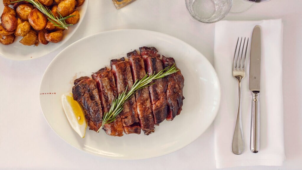 Ein geschnittenes Stück Fleisch liegt auf einem weißen Teller, daneben befindet Besteck sowie eine Schüssel mit Kartoffeln. Ein geschnittenes Stück Fleisch liegt auf einem weißen Teller, daneben befindet Besteck sowie eine Schüssel mit Kartoffeln.