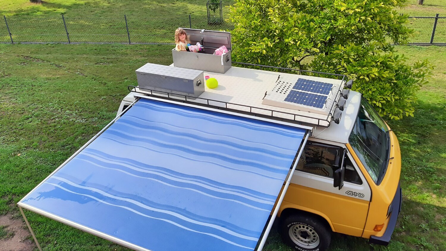 Blick von oben auf einen VW Bus mit Markise und einem Kind in einer Dachbox