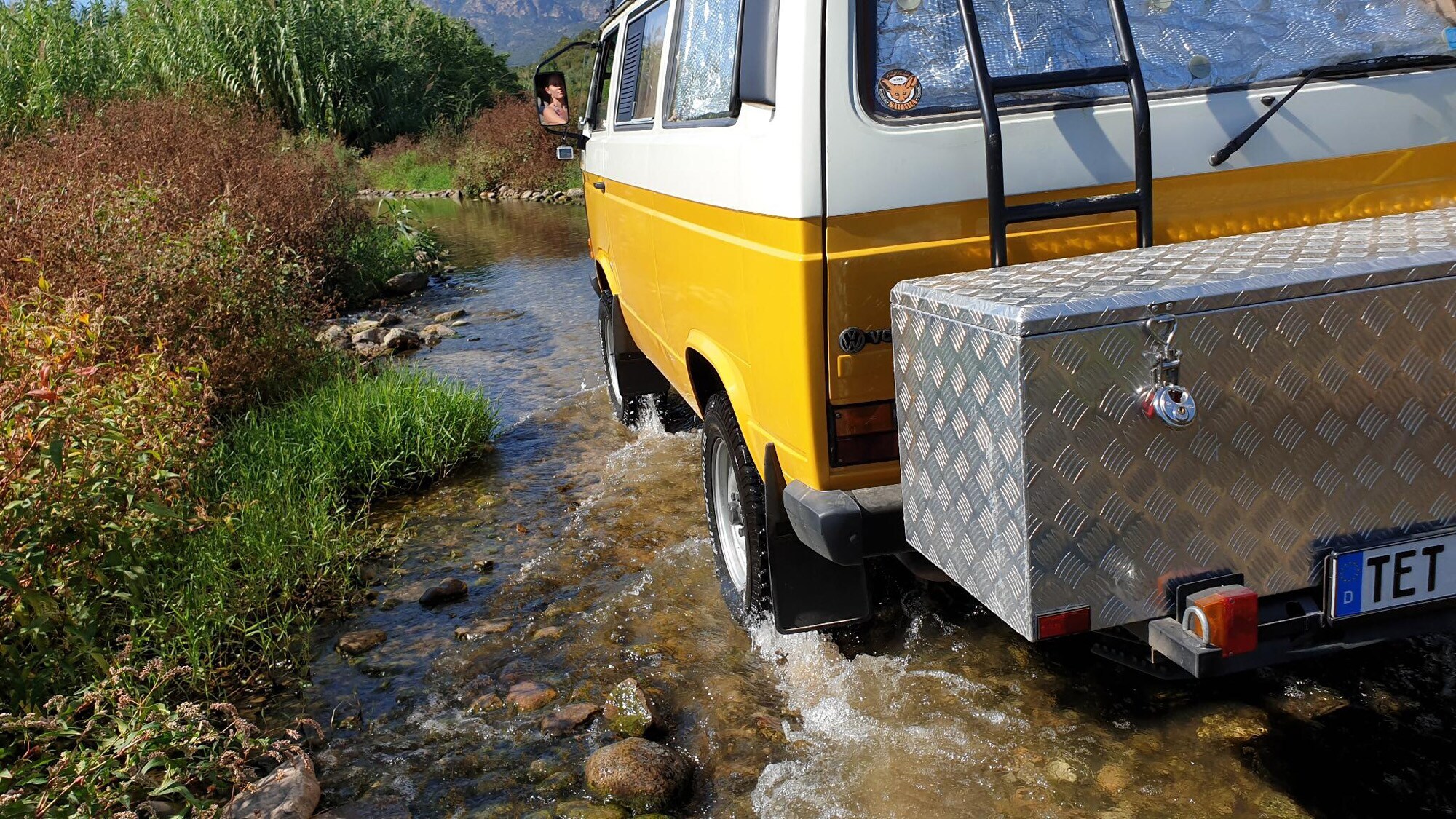 Heckansicht eines gelben VW Bus mit großer Heckbox, der durch ein Flussbett fährt Heckansicht eines gelben VW Bus mit großer Heckbox, der durch ein Flussbett fährt