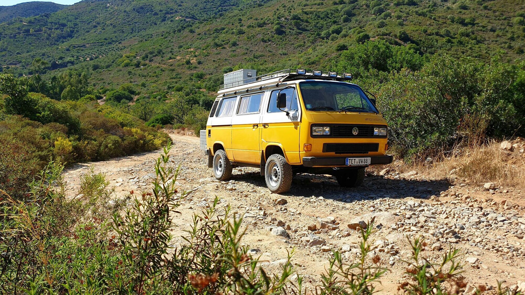 Ein gelber VW Bus auf einer Schotterstraße in einer bergigen Landschaft