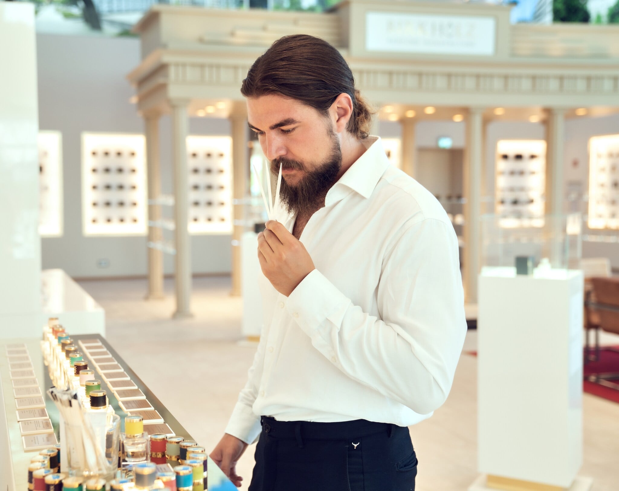 Ein Mann mit Vollbart in einem weißen Hemd riecht an drei Duft-Teststreifen im Showroom einer Parfümmanufaktur Ein Mann mit Vollbart in einem weißen Hemd riecht an drei Duft-Teststreifen im Showroom einer Parfümmanufaktur