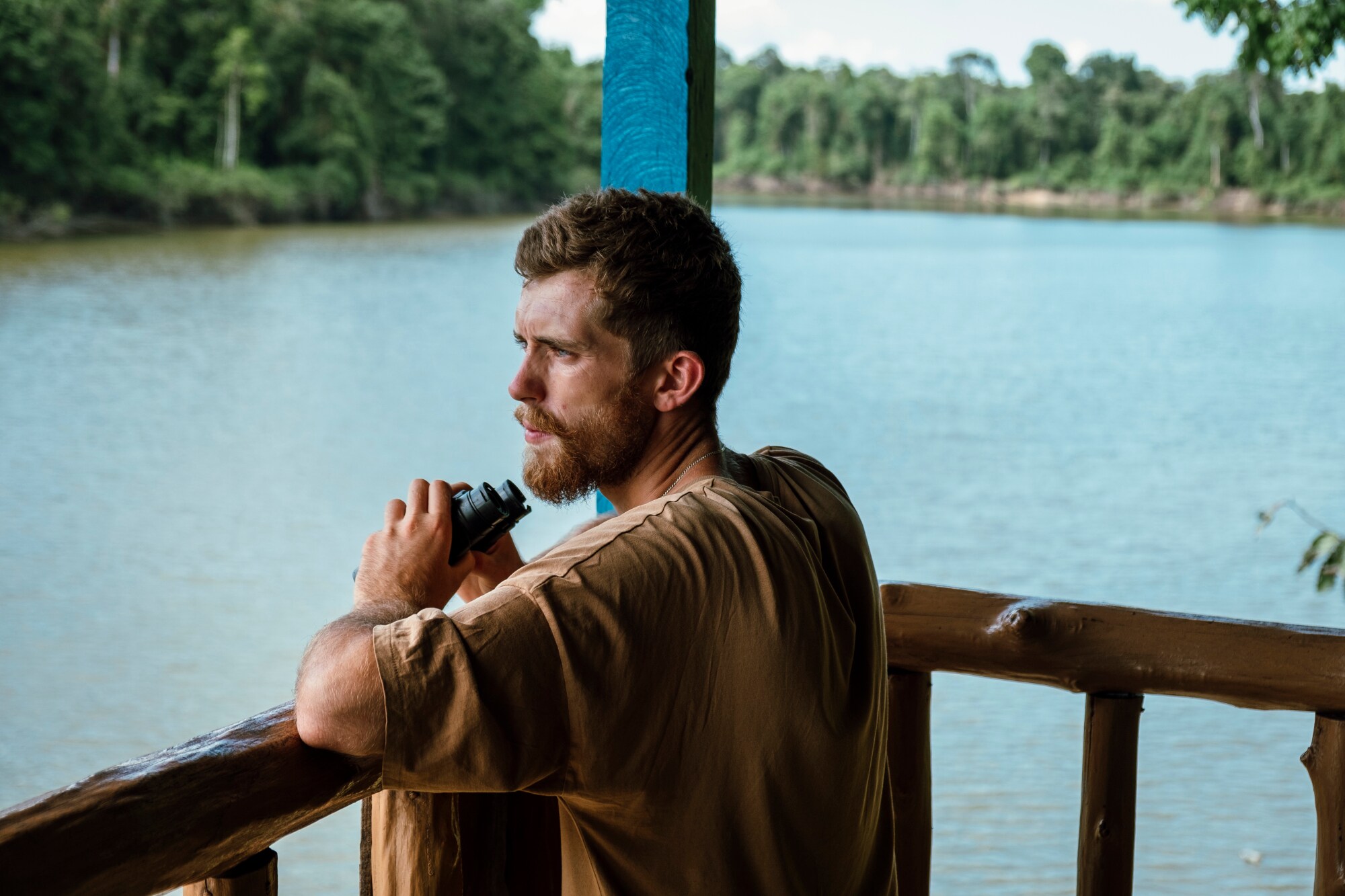 Ein junger Mann mit Bart steht an der Reling eines Hausbootes auf einem Fluss und schaut in den Regenwald, in der Hand hält er ein Fernglas. Ein junger Mann mit Bart steht an der Reling eines Hausbootes auf einem Fluss und schaut in den Regenwald, in der Hand hält er ein Fernglas.