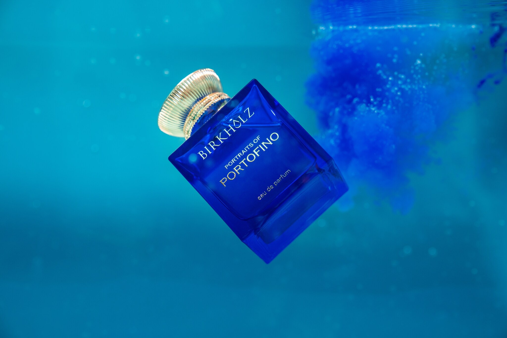 Ein eleganter, quadratischer Parfumflakon aus blauem Glas der Marke Birkholz mit goldenem Verschluss schwebt in blauem Wasser Ein eleganter, quadratischer Parfumflakon aus blauem Glas der Marke Birkholz mit goldenem Verschluss schwebt in blauem Wasser