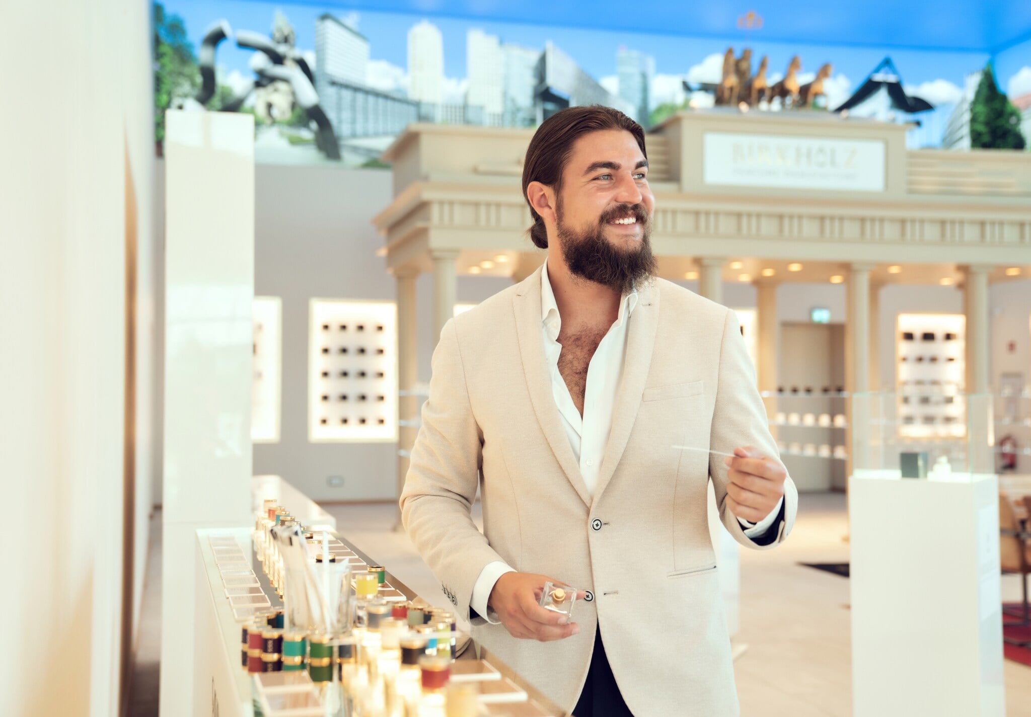 Ein Mann mit Vollbart in einem weißen Jackett steht im hellen Verkaufsraum einer Parfümmanufaktur und testet ein Parfüm mit Duft-Teststreifen