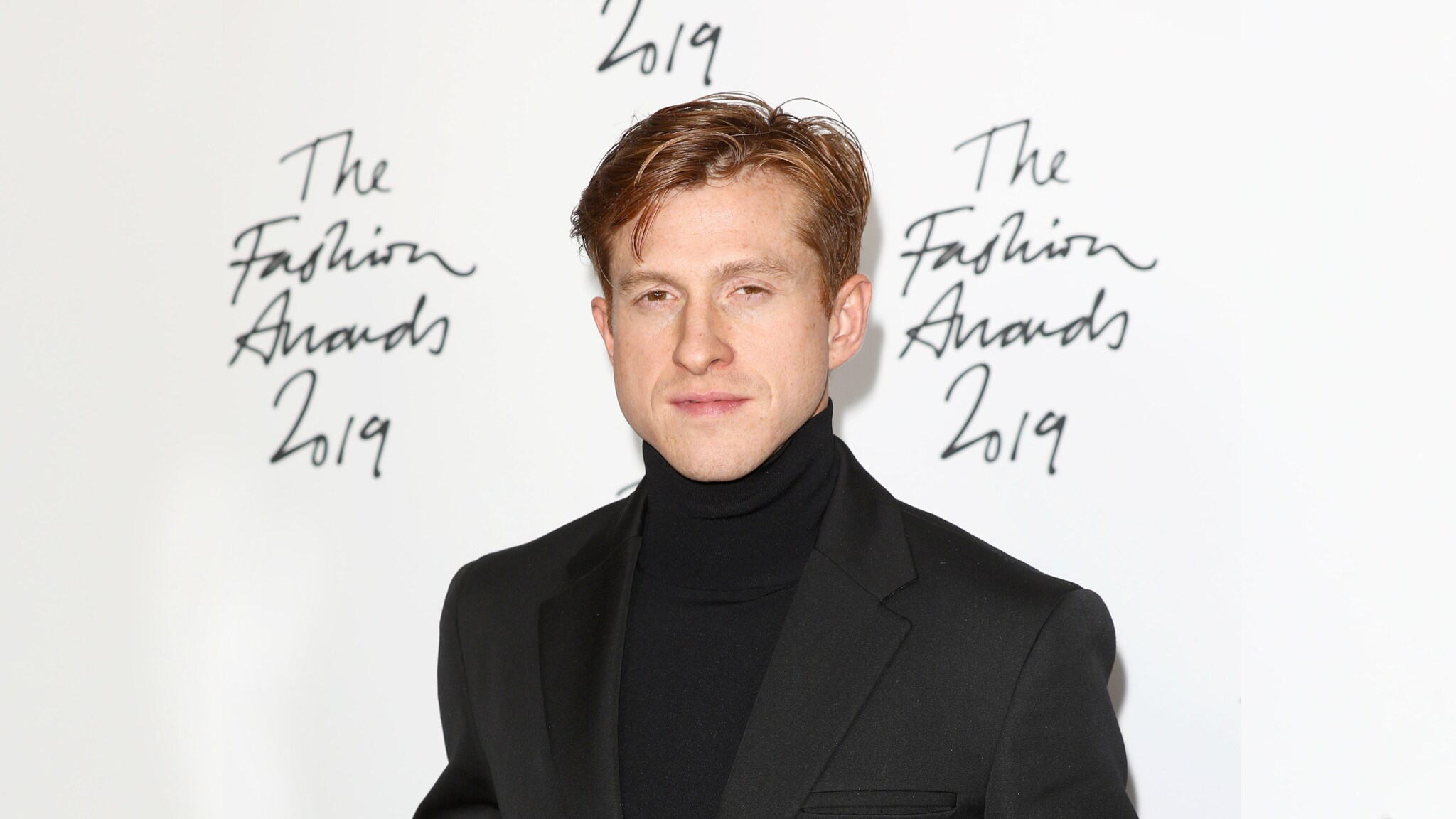 Ein Mann im Anzug posiert vor einer Wand, auf der ein Schriftzug zu lesen ist.