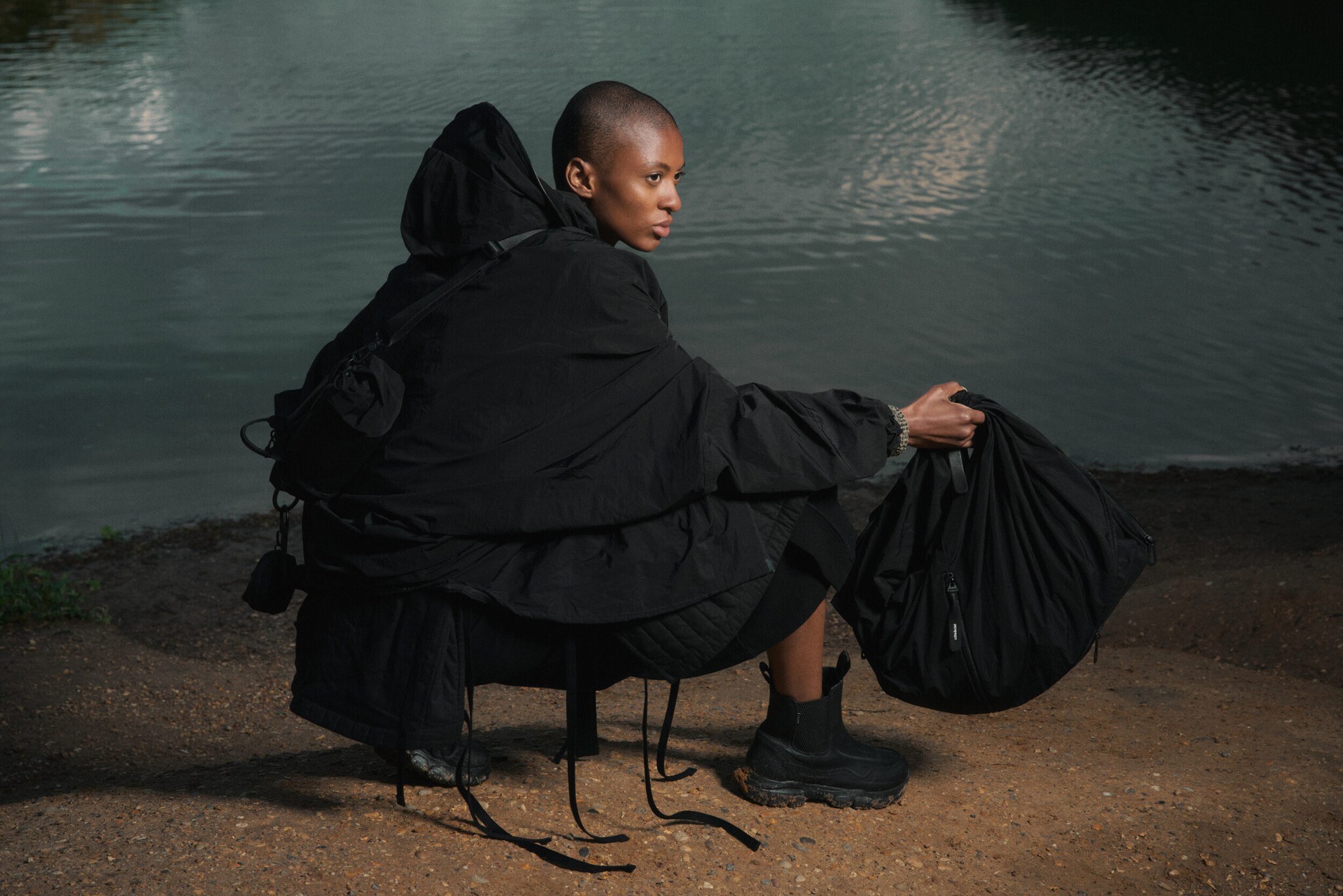 In Schwarz gekleidete Frau mit Taschen von côte&ciel in der Hand und auf dem Rücken vor einer Kulisse am See