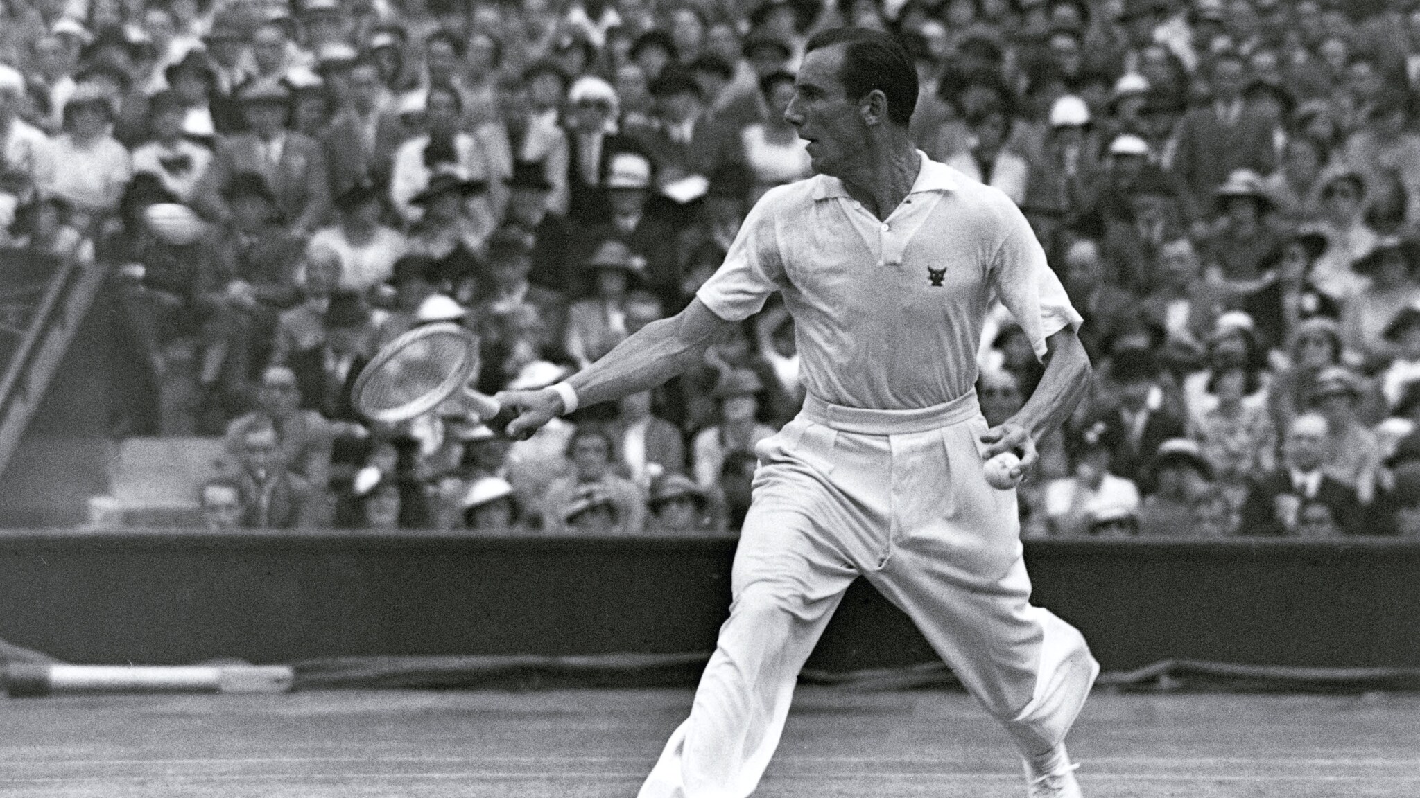 Schwarzweißaufnahme von Fred Perry in weißer Sportkleidung bei einem Tennisspiel vor Publikum Schwarzweißaufnahme von Fred Perry in weißer Sportkleidung bei einem Tennisspiel vor Publikum