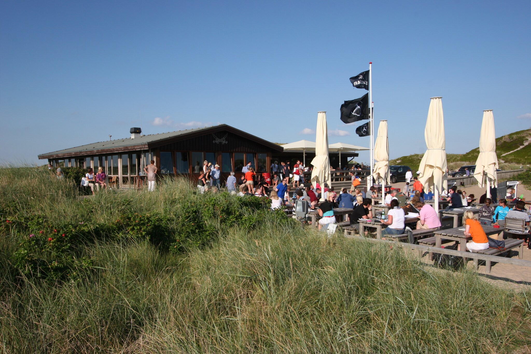 Viele Menschen sitzen an Holztischen und -bänken im Freien vor einem Haus.