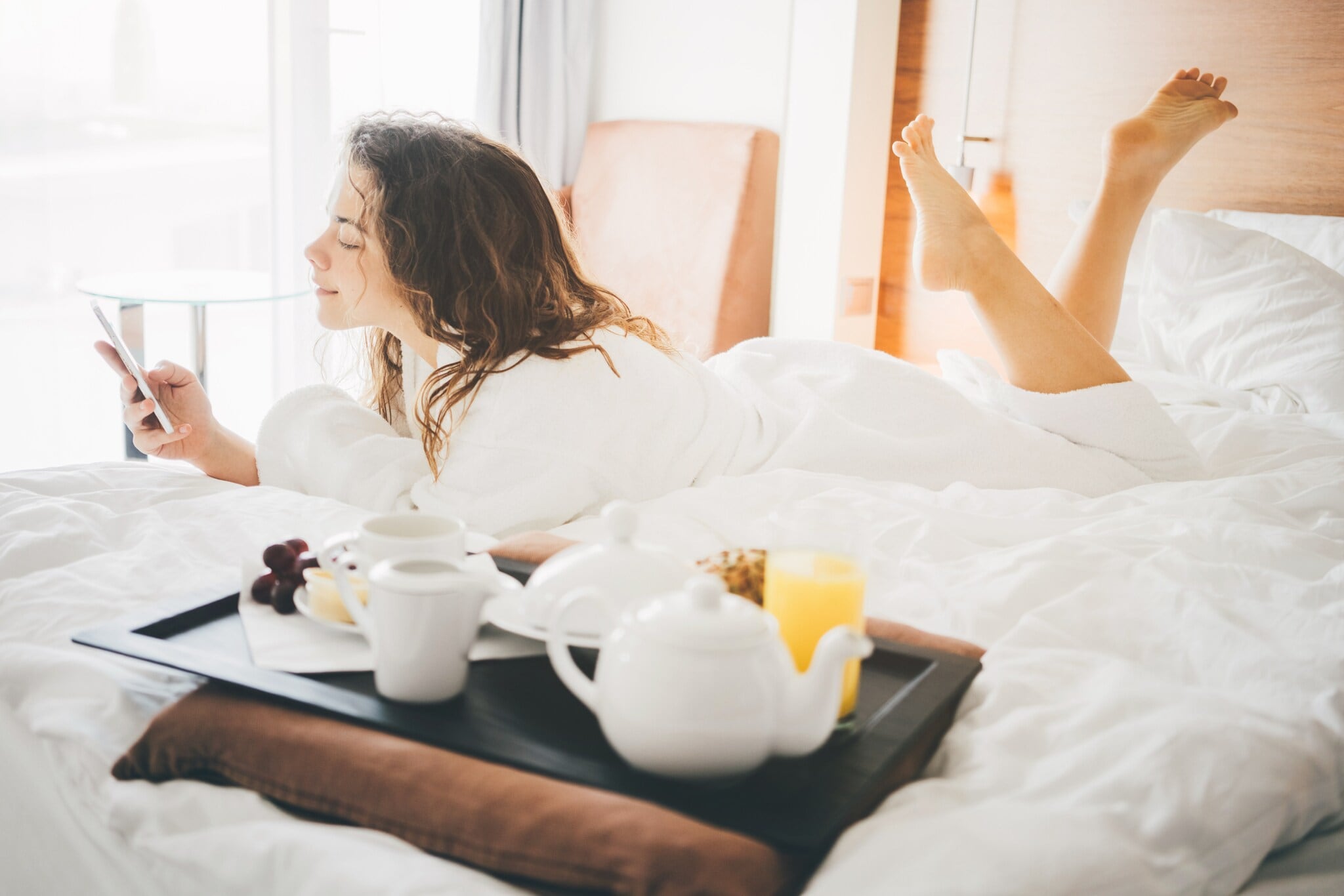 Eine Frau liegt im Bademandel auf einem Bett in ihrem Hotelzimmer. In der Hand hält sie ein Smartphone, neben ihr steht ein Tablett mit Frühstück. Eine Frau liegt im Bademandel auf einem Bett in ihrem Hotelzimmer. In der Hand hält sie ein Smartphone, neben ihr steht ein Tablett mit Frühstück.