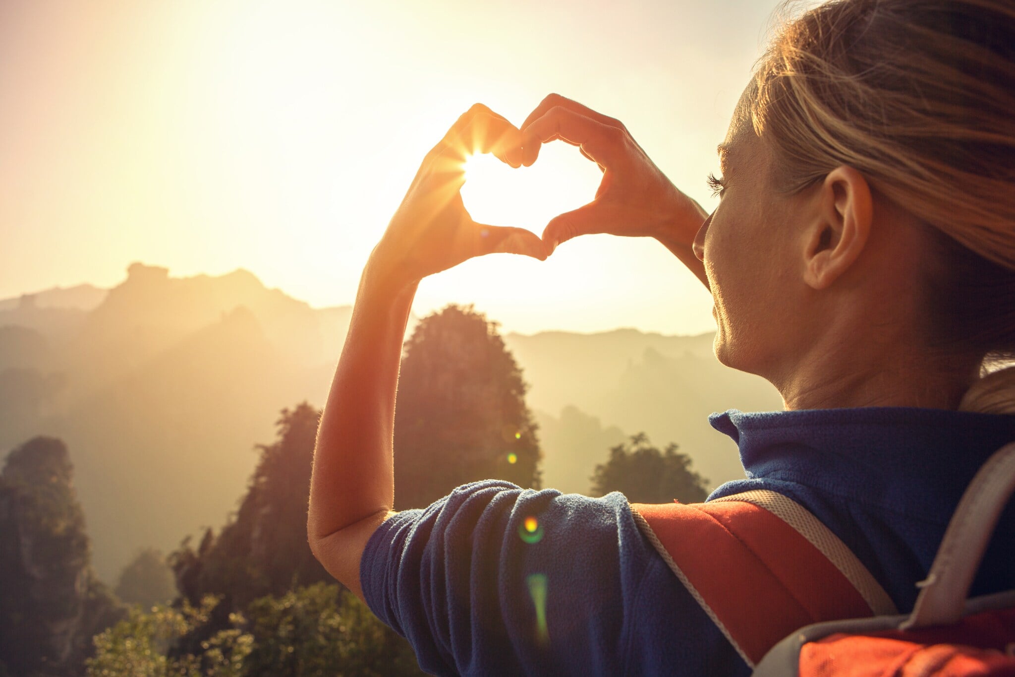 Eine junge Frau blickt in die Natur und formt ihre Hände zu einem Herz, durch das die Sonne scheint Eine junge Frau blickt in die Natur und formt ihre Hände zu einem Herz, durch das die Sonne scheint