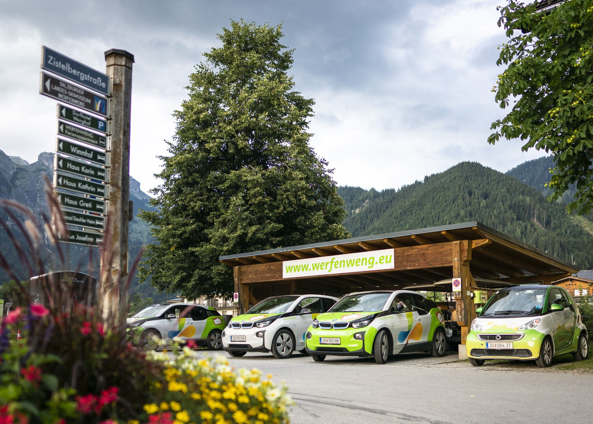 Mehrere E-Autos stehen unter einem Carport, während im Hintergrund begrünte Berge zu erkennen sind.