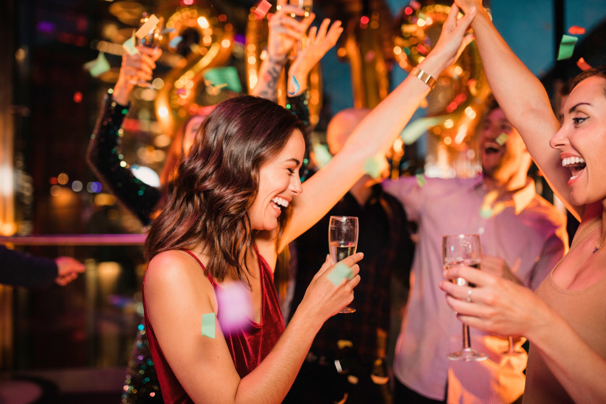 Zwei Frauen mit Sektgläsern in der Hand feiern auf einer Party und lachen, im Hintergrund sind weitere Gäst:innen zu sehen.