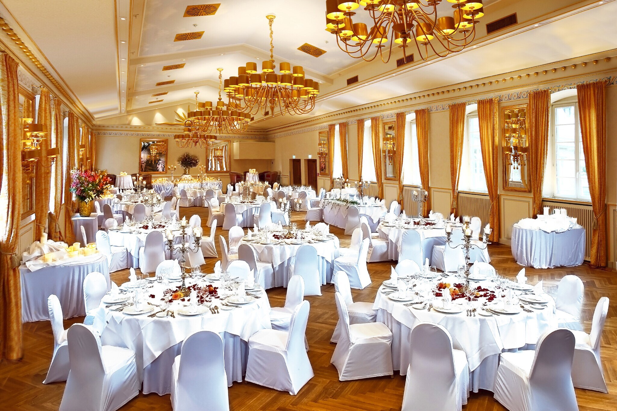 Stilvoll eingedeckte runde Tische mit weißen Stühlen in einem großen Festsaal mit Fischgrätenparkett. Stilvoll eingedeckte runde Tische mit weißen Stühlen in einem großen Festsaal mit Fischgrätenparkett.