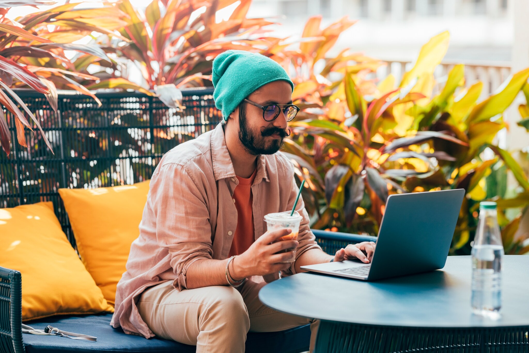 Ein junger Mann mit Bart und Mütze sitzt im Freien am Laptop, in der Hand hält er ein Getränk. Ein junger Mann mit Bart und Mütze sitzt im Freien am Laptop, in der Hand hält er ein Getränk.