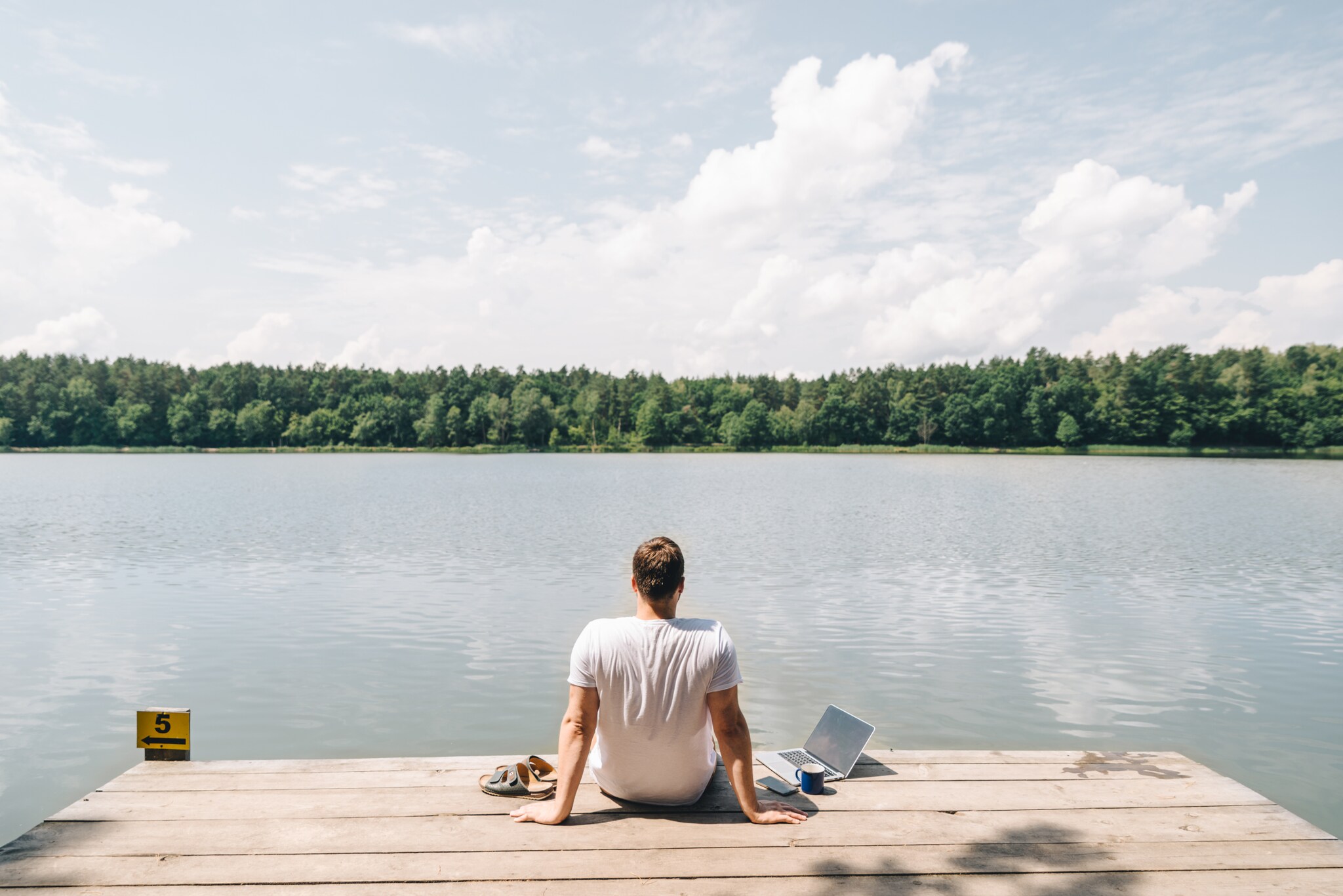 Ein Mann sitzt auf einem Steg am See und schaut aufs Wasser, neben sich ein Laptop.